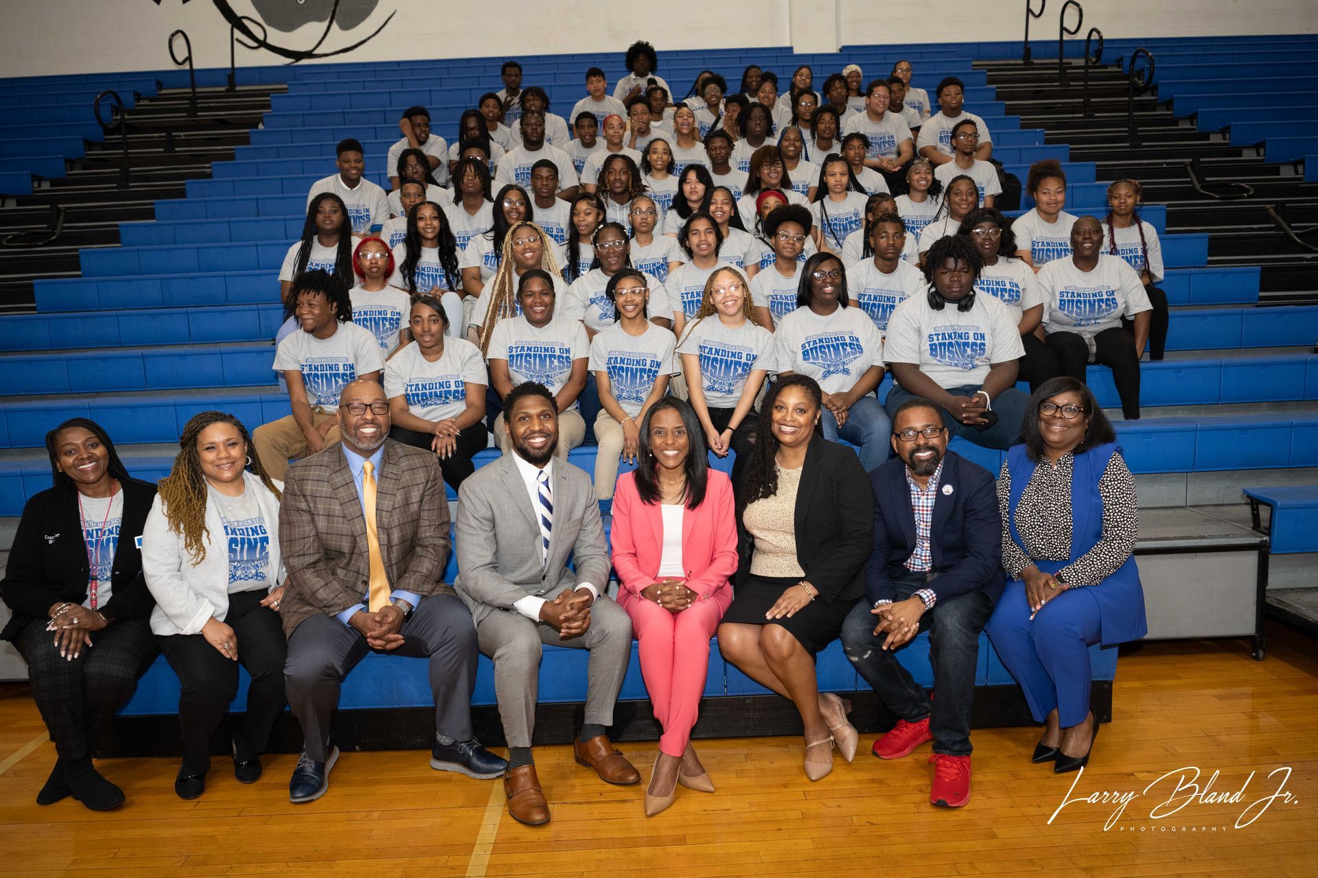 Group photo in a gymnasium. People wearing matching shirts sit on bleachers. Adults in business attire sit in front row.