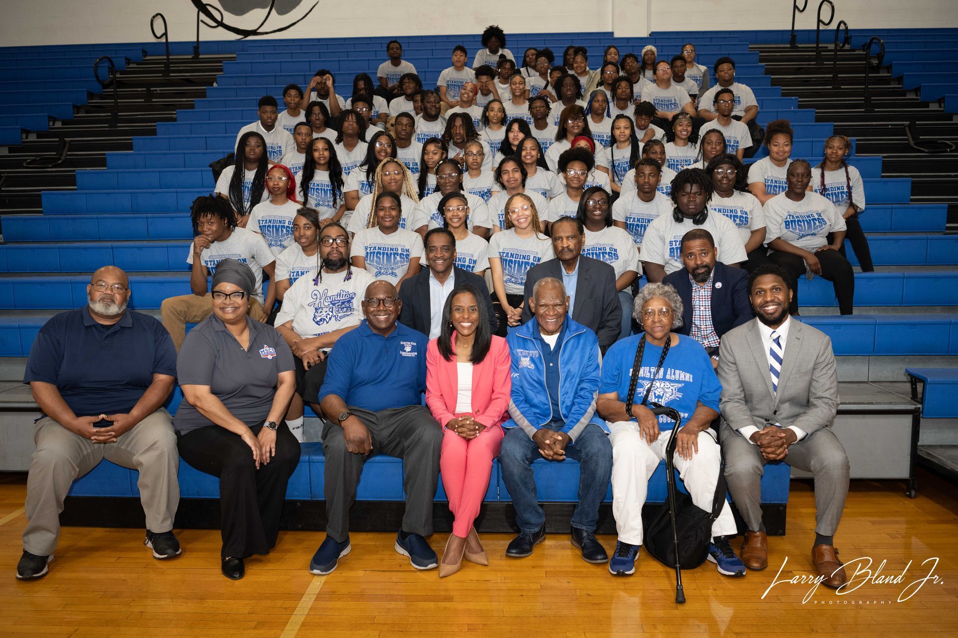 Group photo: people in a gymnasium with blue bleachers. People in the front row are seated. Some wear blue and white shirts.