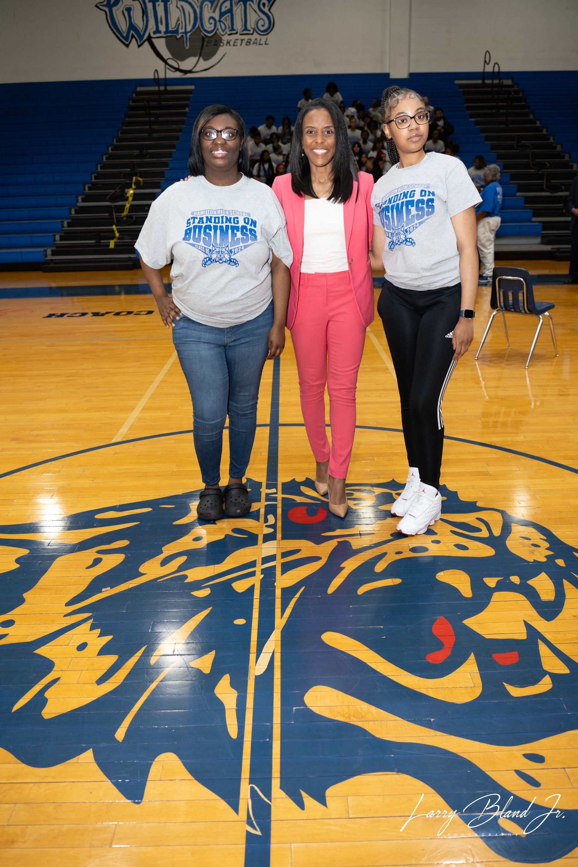 Three people stand on a basketball court. One in a pink suit, two in t-shirts and jeans. A Wildcats logo is on the floor.