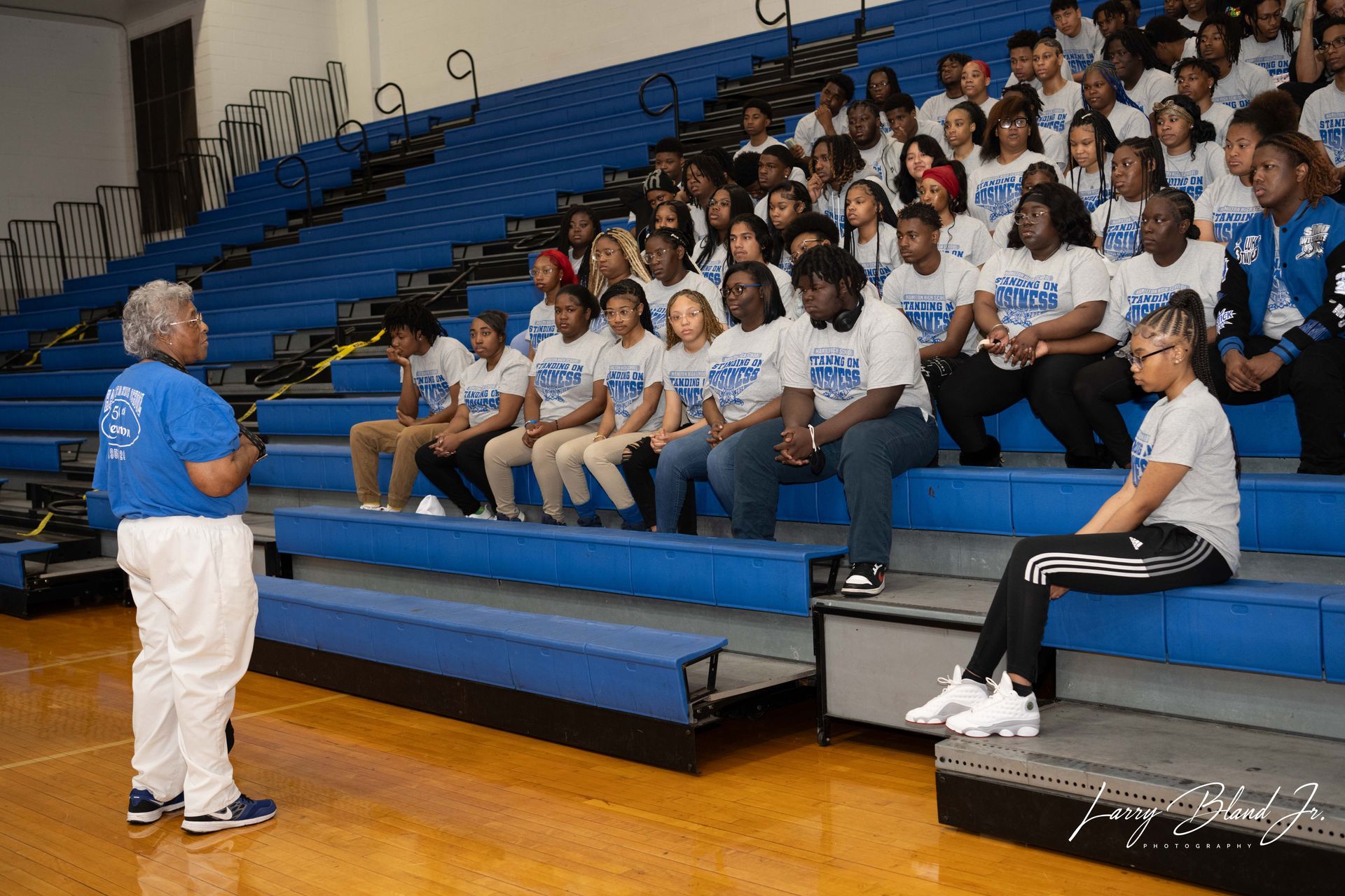 A woman speaks to a group of students in a blue and white gym. Students are seated on bleachers.