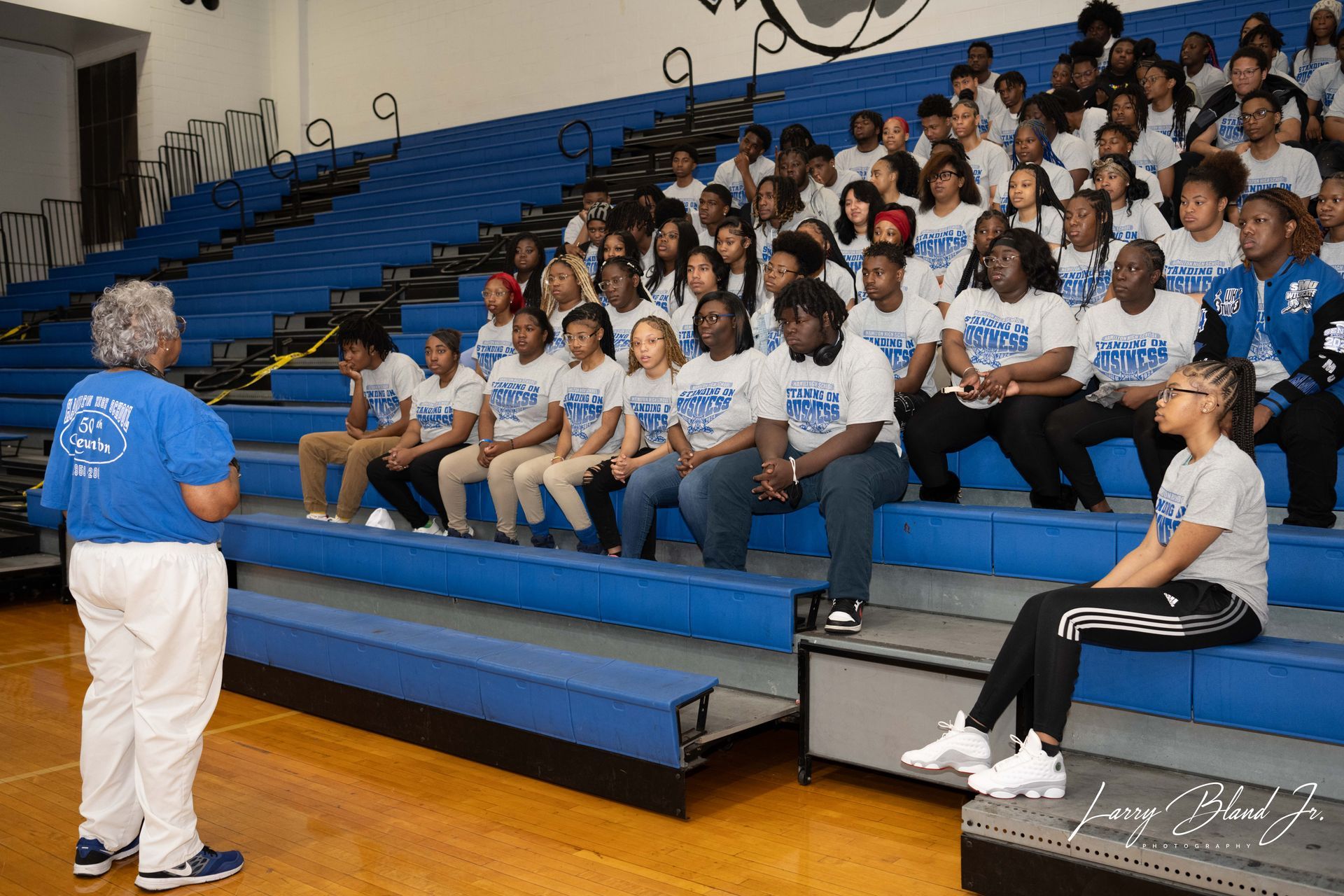 Woman speaks to a seated group in a blue-seated auditorium. They wear matching t-shirts; some smile.