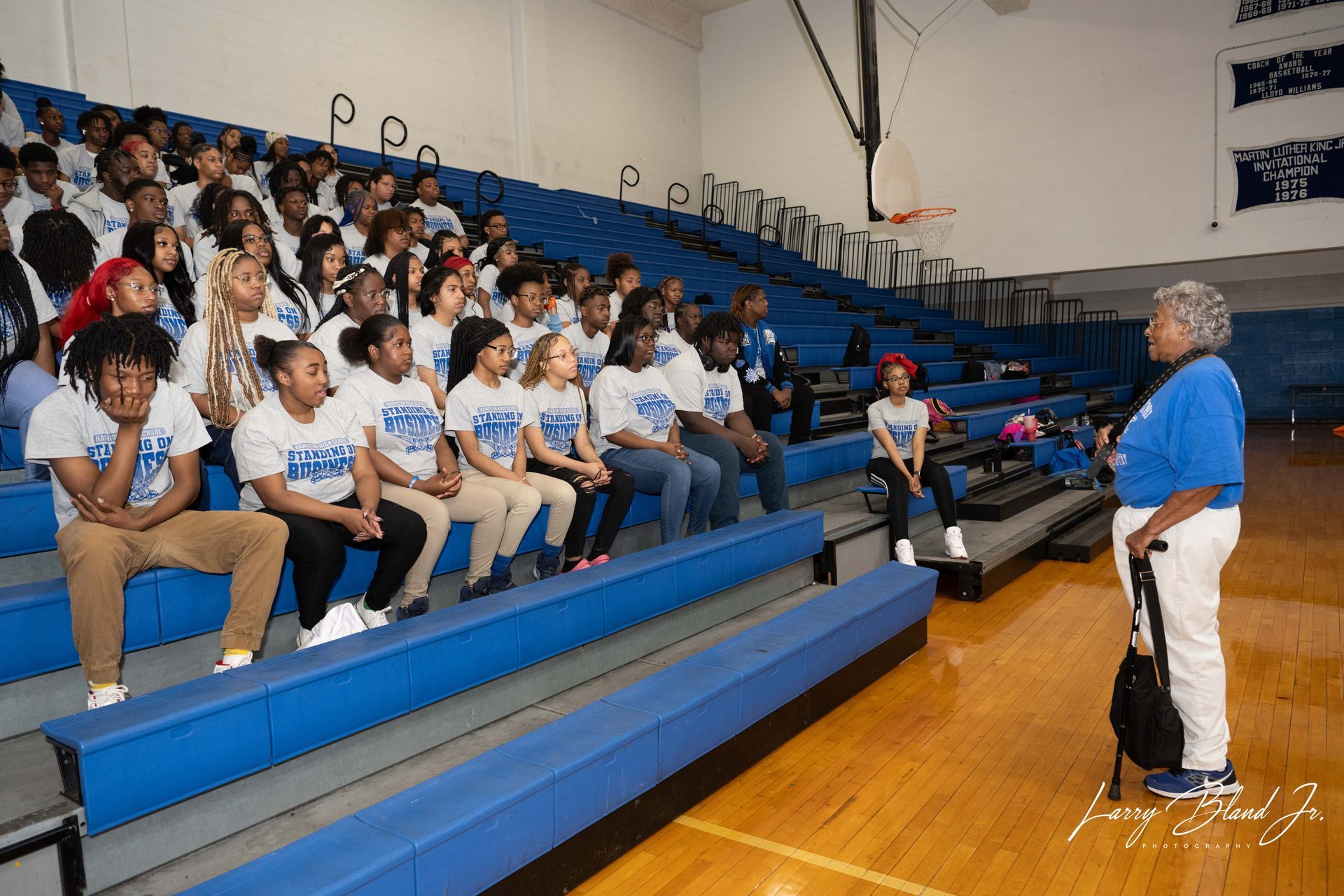 Audience in bleachers listens to a speaker in a gymnasium. Blue and white colors.