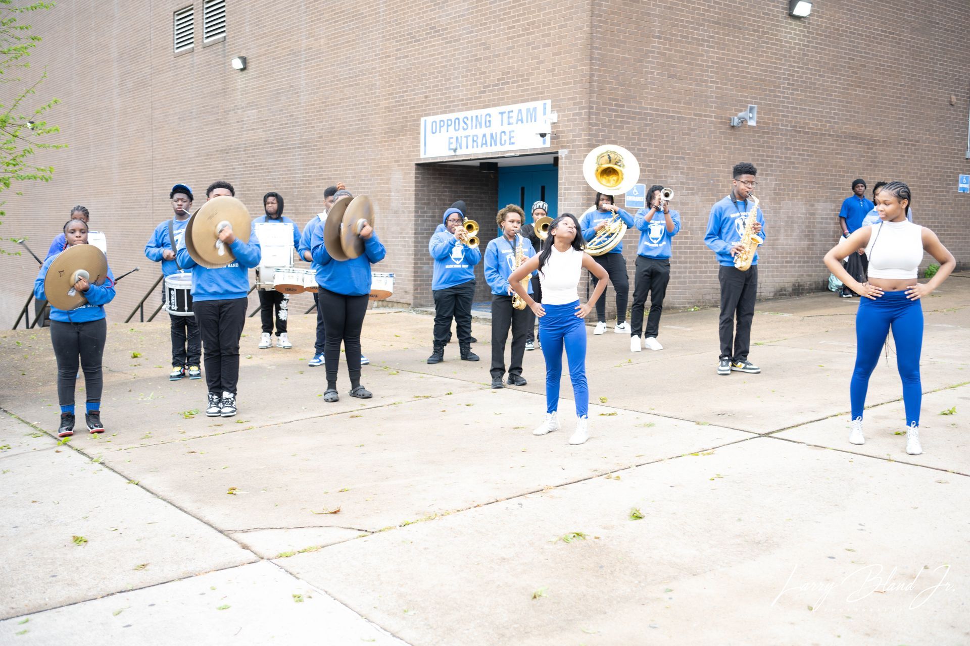 Band in blue uniforms performing outside a building, with two dancers in front.