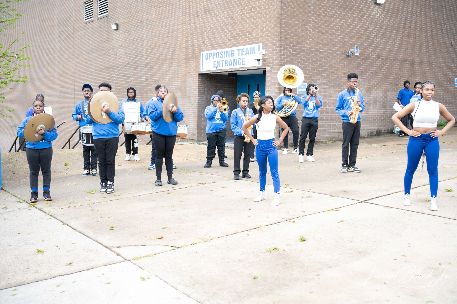 A marching band in blue uniforms performs outside a building. Two dancers pose.
