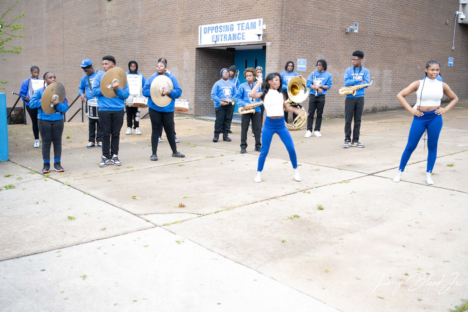 A band in blue outfits performs outdoors; a dancer in the center, and a building with 