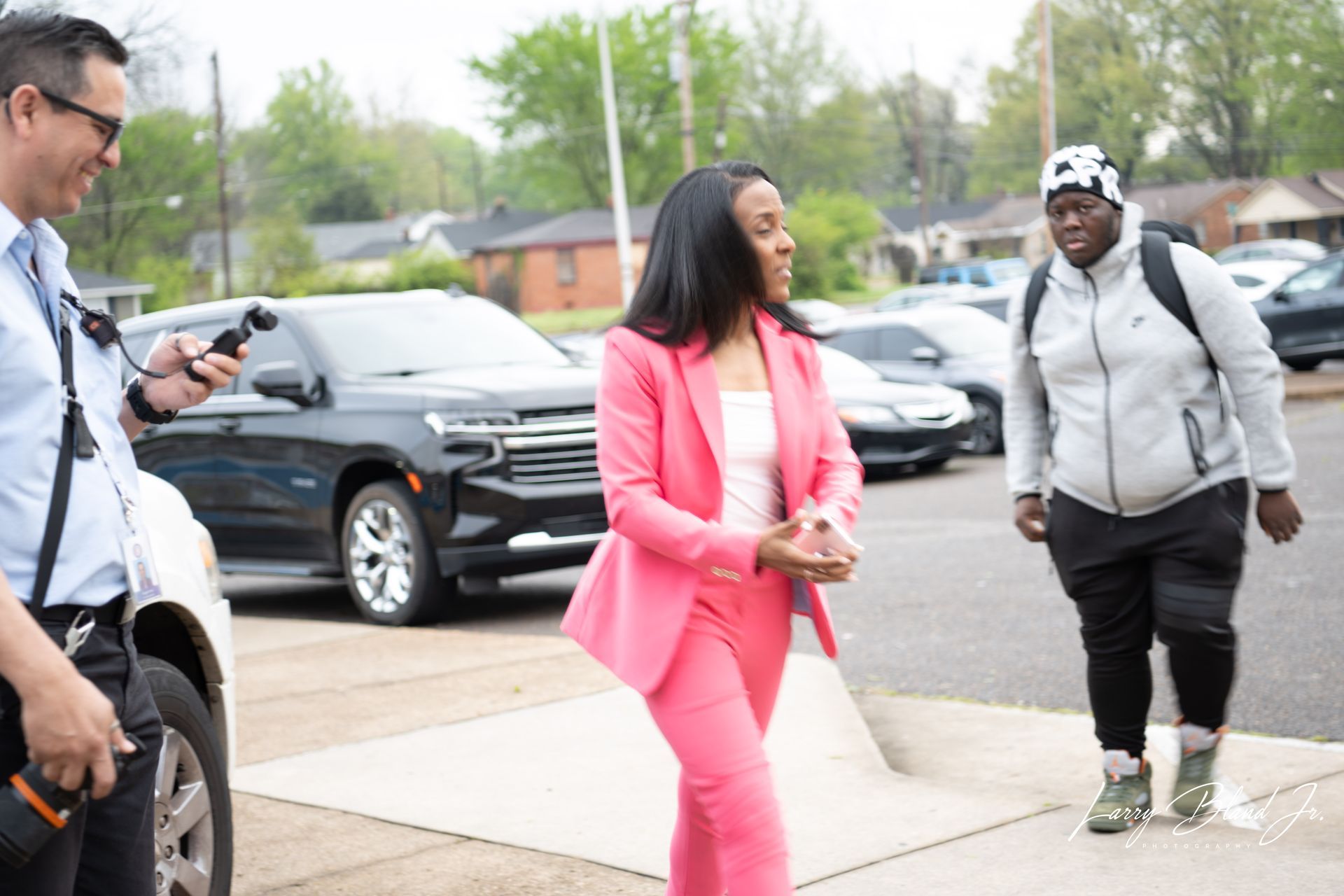 Woman in pink suit exits building, flanked by man with phone and person with backpack.