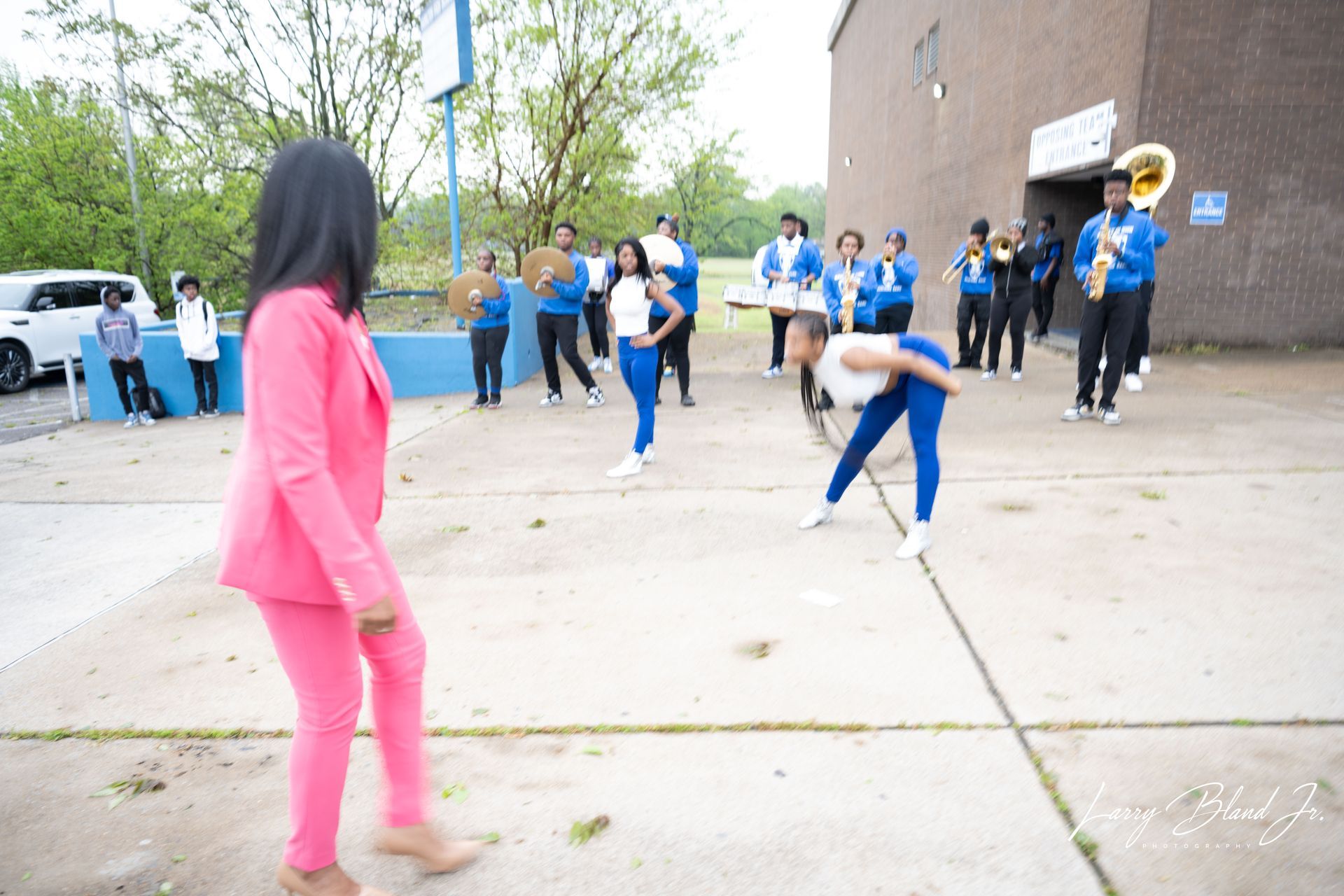 A person in pink suit watches a marching band in blue and gold uniforms outside a brick building.