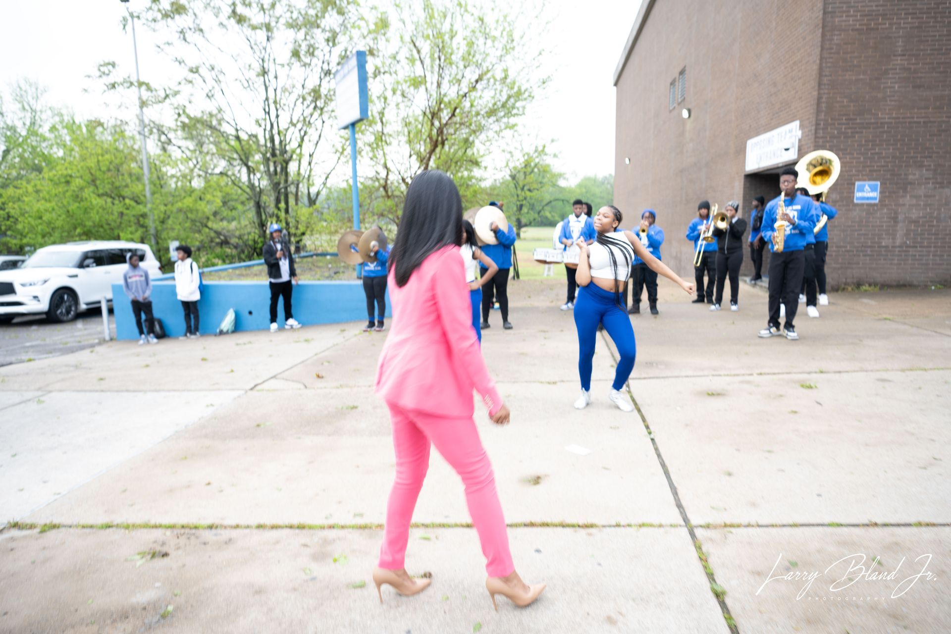 Woman in pink suit faces a marching band in blue and gold uniforms outside a building.