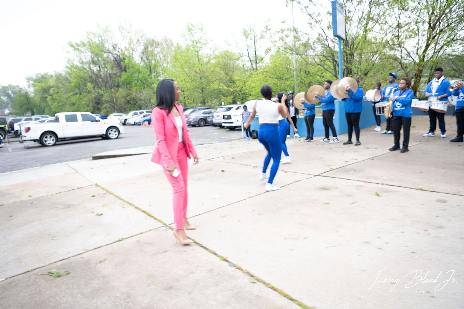Woman in pink suit watches a marching band in blue uniforms perform outside.