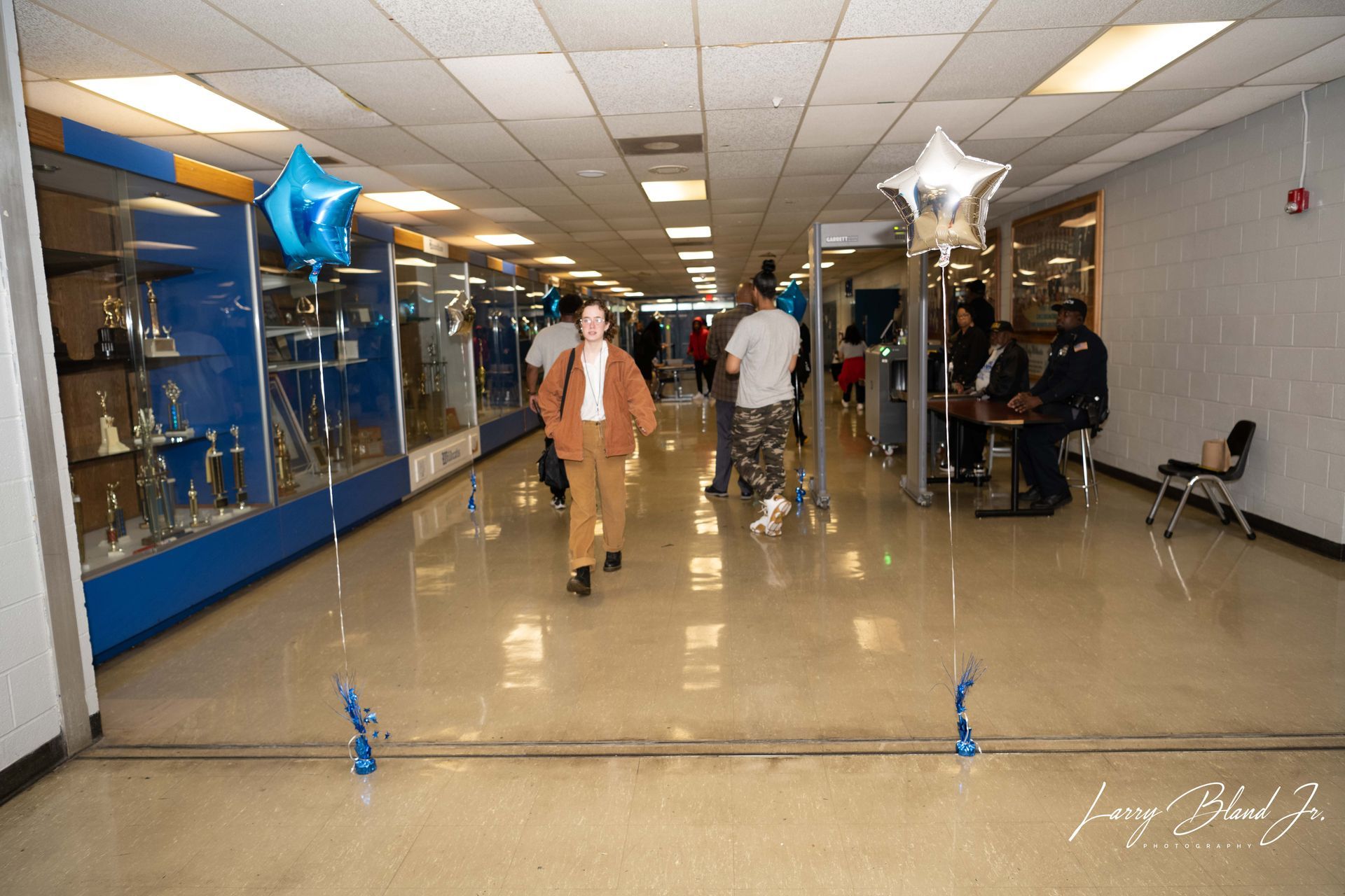 School hallway with students and balloons. A person walks toward the camera. Display cases and tables are set up.