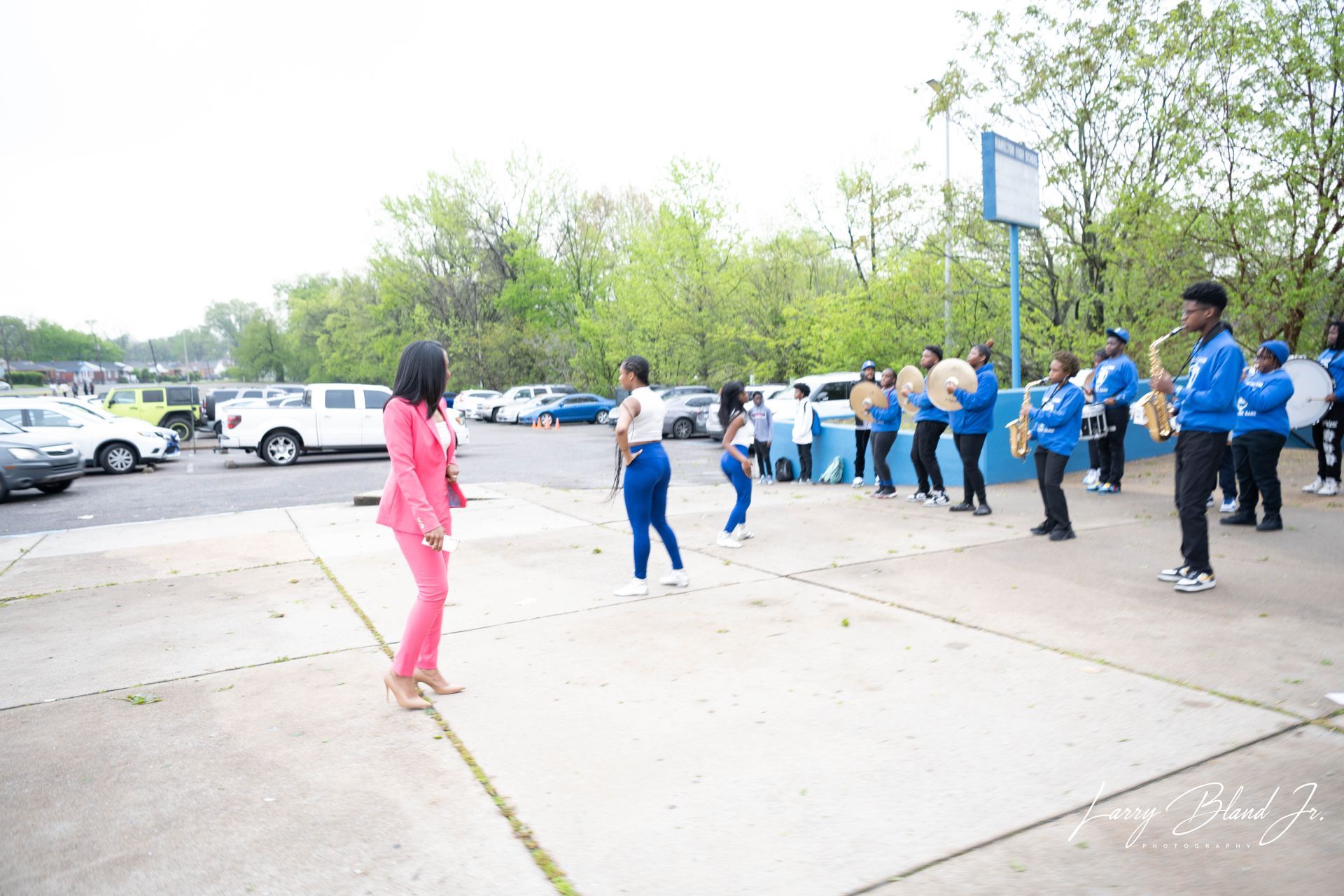 A marching band in blue uniforms performs outside. A woman in pink stands nearby.