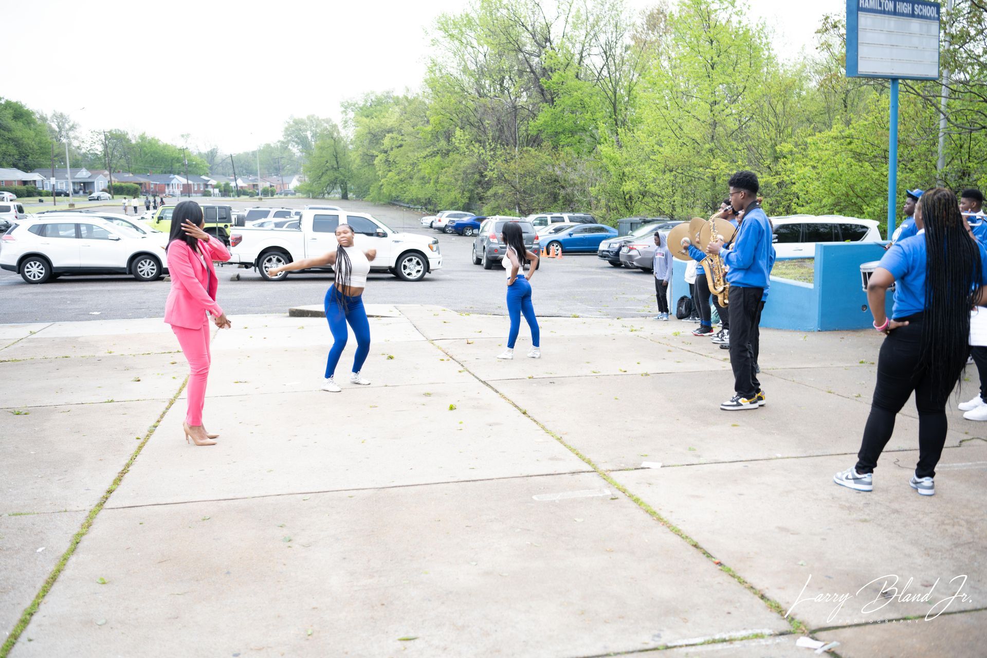 Group of people outside; some dancing, others watching. Building and parked cars in background.
