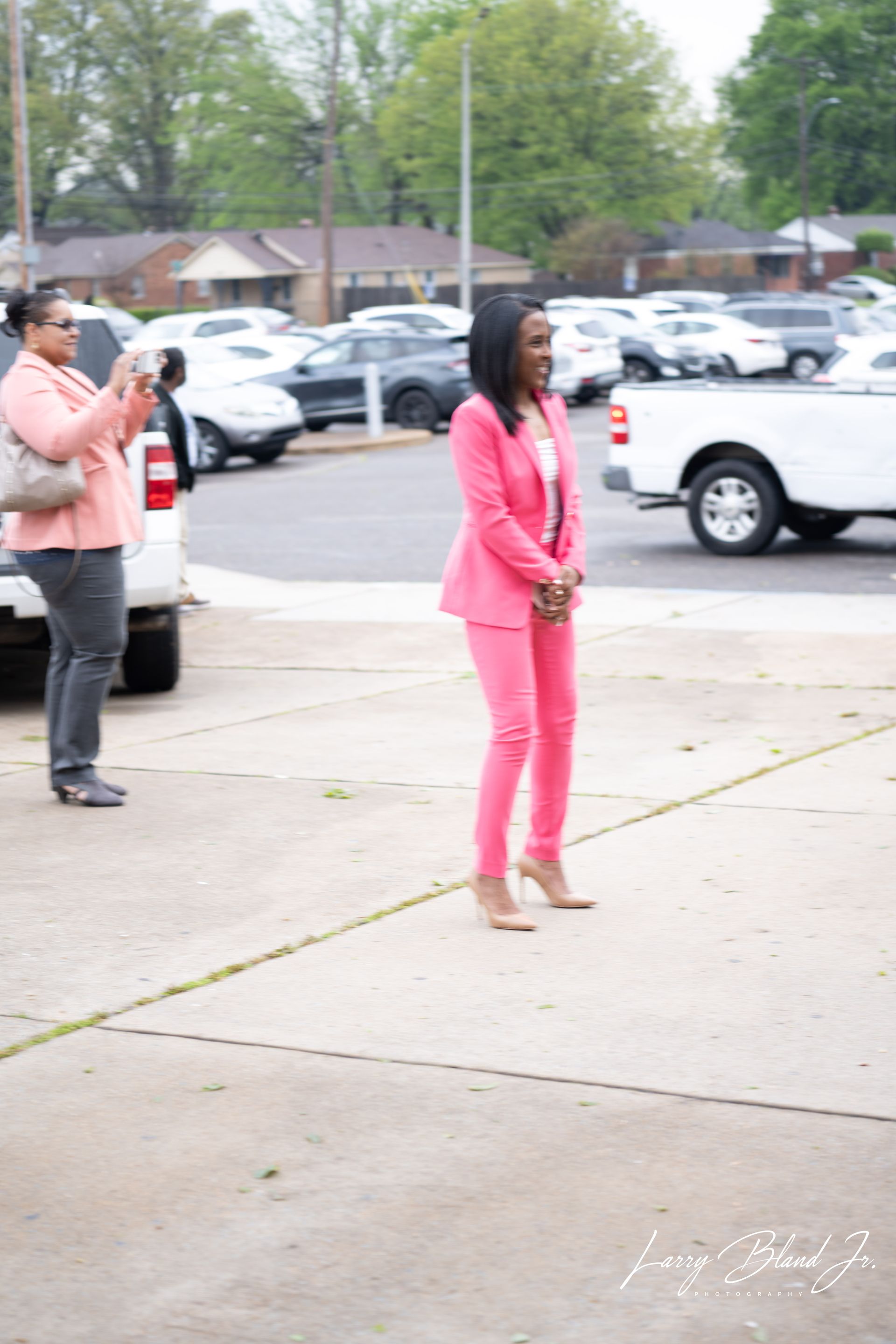 Woman in pink suit, standing outside with another person taking a photo. White cars and a building in the background.
