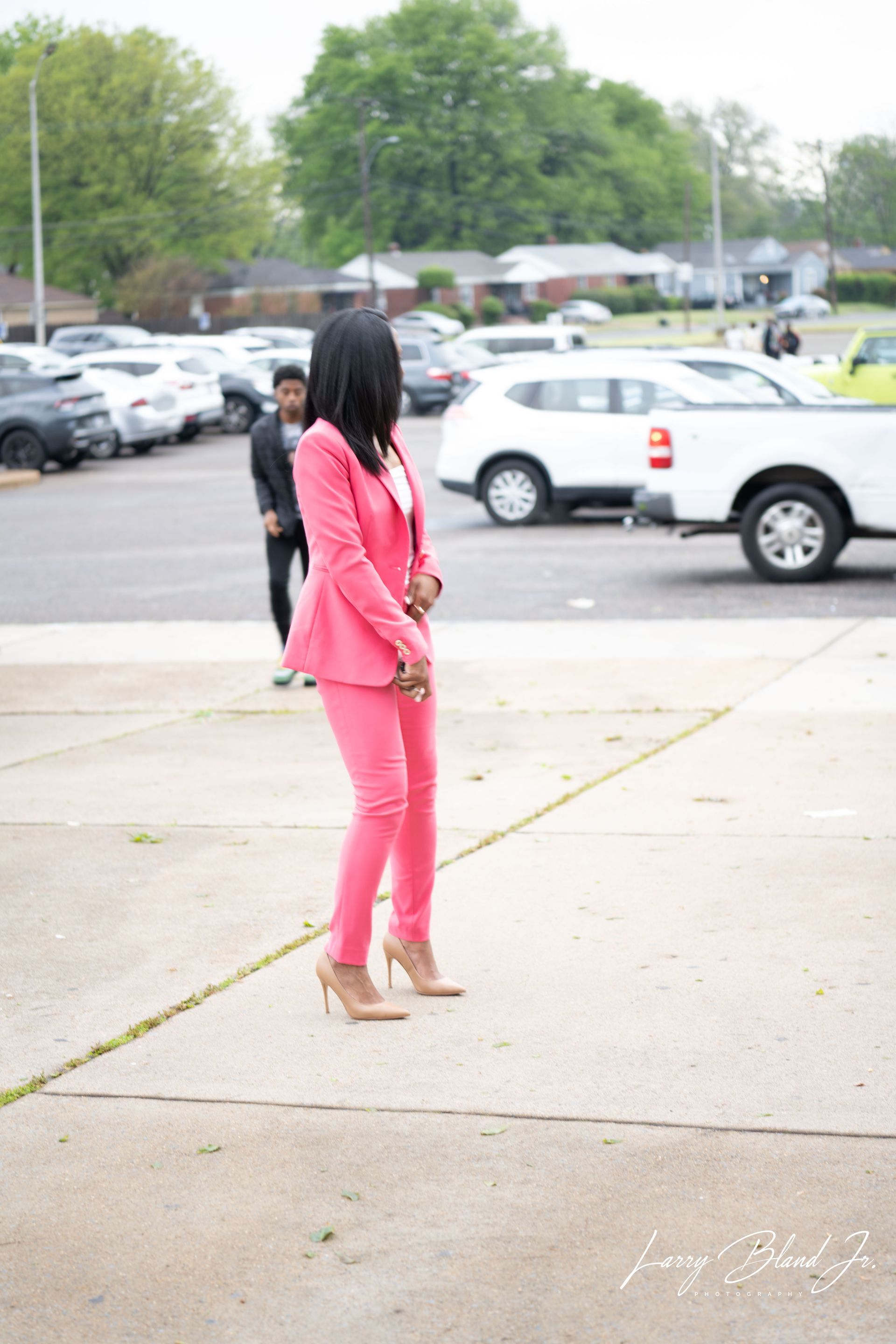 Woman in pink suit and heels stands outdoors; cars and building in the background.