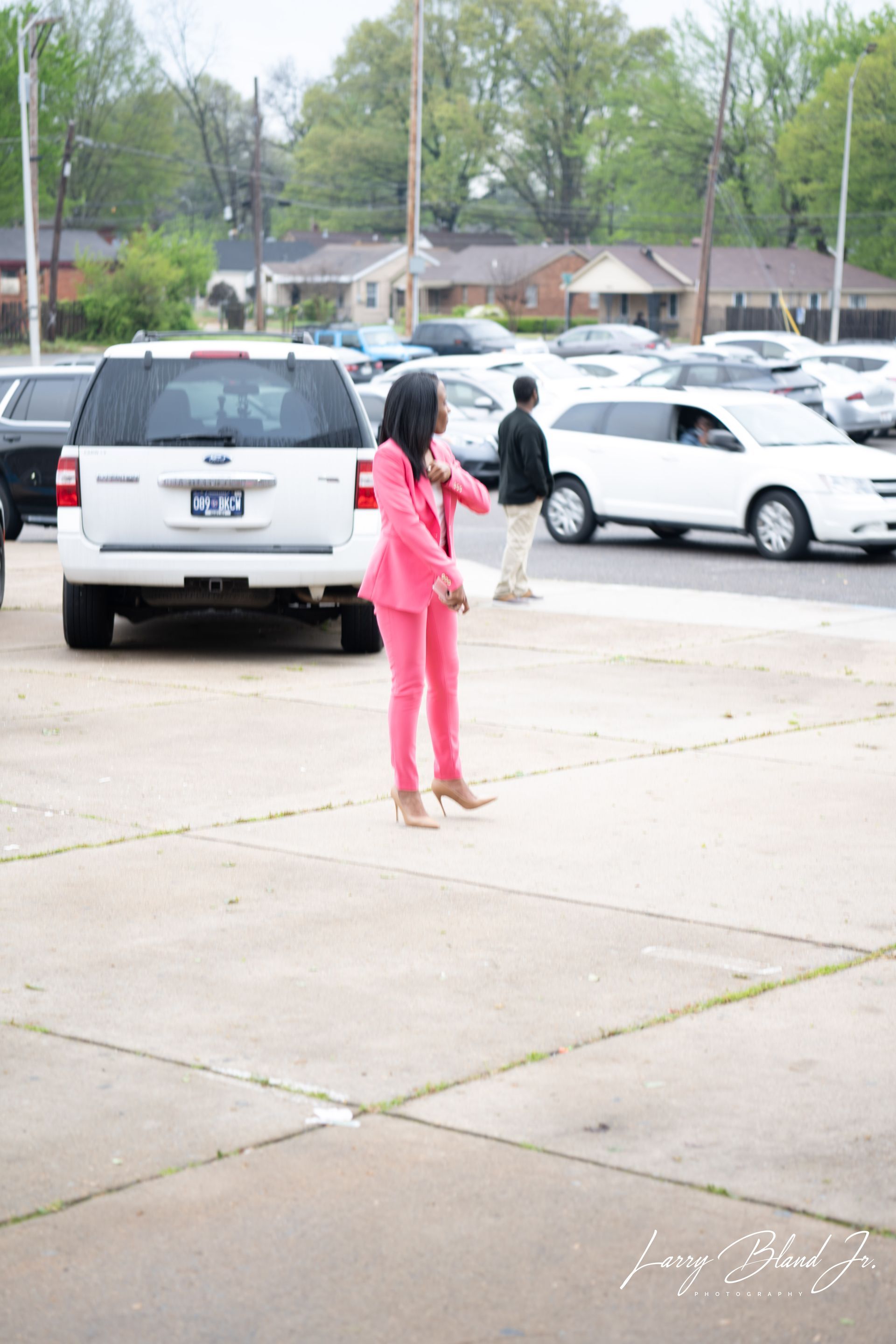 Woman in pink suit, dancing in parking lot. Cars and buildings in background.