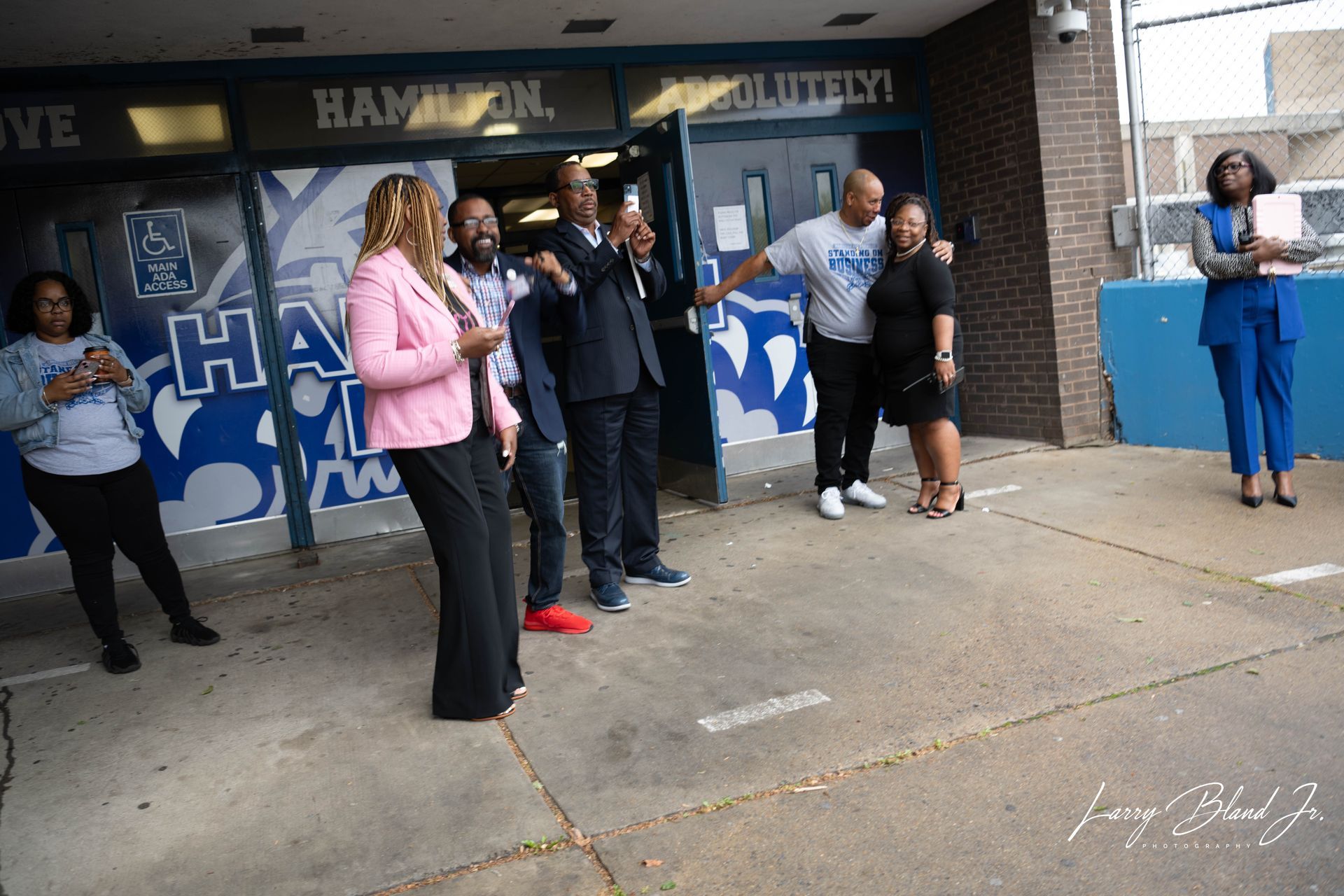 People gather outside a school with blue and white accents; some hold phones, smiling.