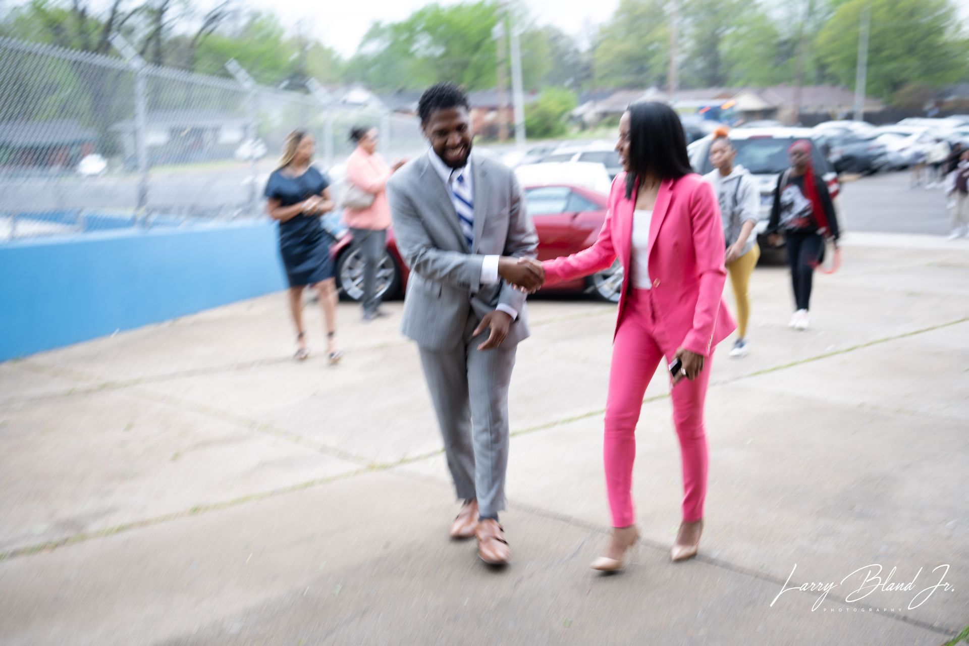 Man and woman in suits shaking hands outside a building, other people nearby.