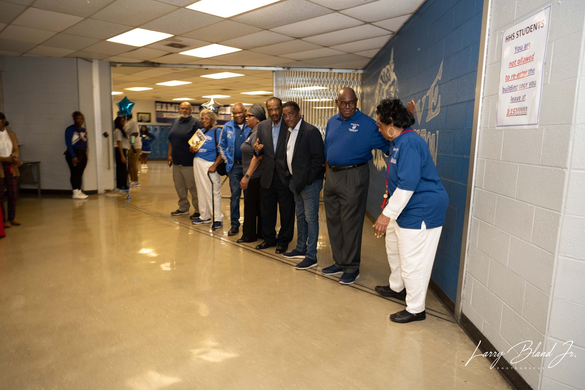 People stand in hallway; some wear blue, near decorated wall.