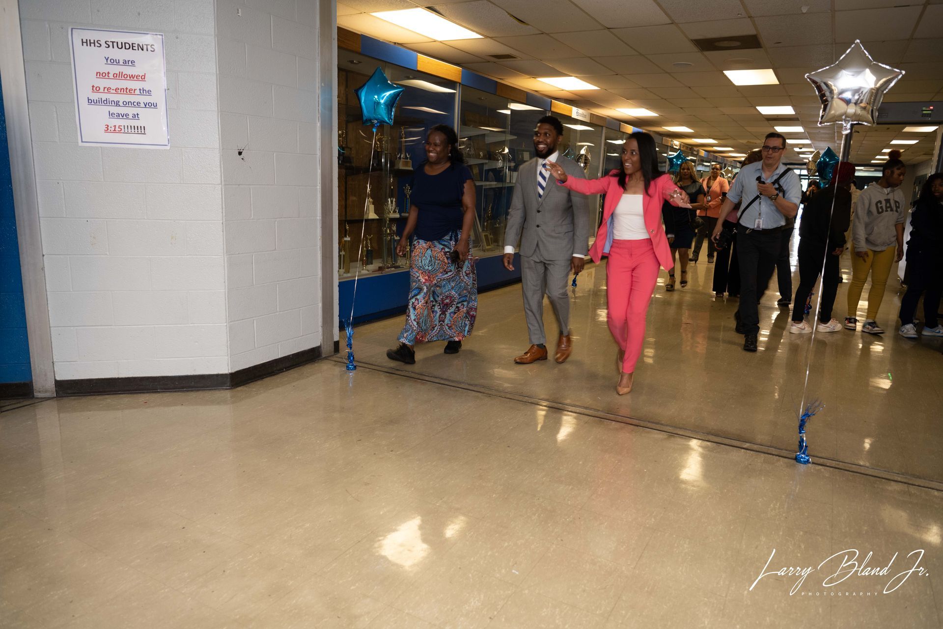People walking in a school hallway, celebrating. One woman in pink suit leads the way, balloons present.