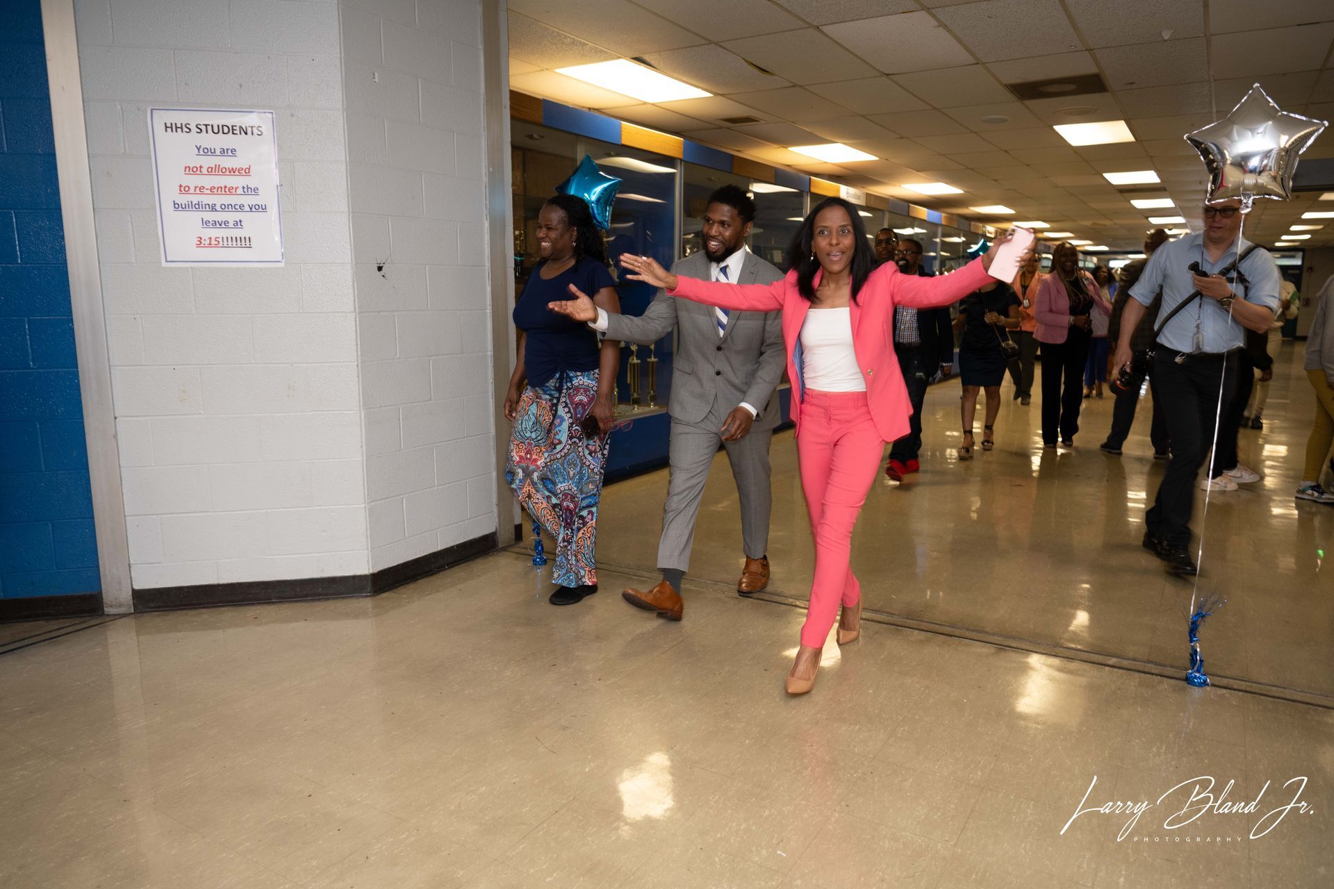 Three people celebrate in a school hallway: woman in pink suit with arms out, man in grey suit, and another woman.