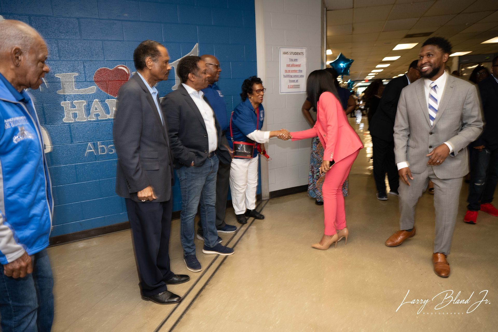People shake hands in a hallway; one person wears a pink suit, blue wall.