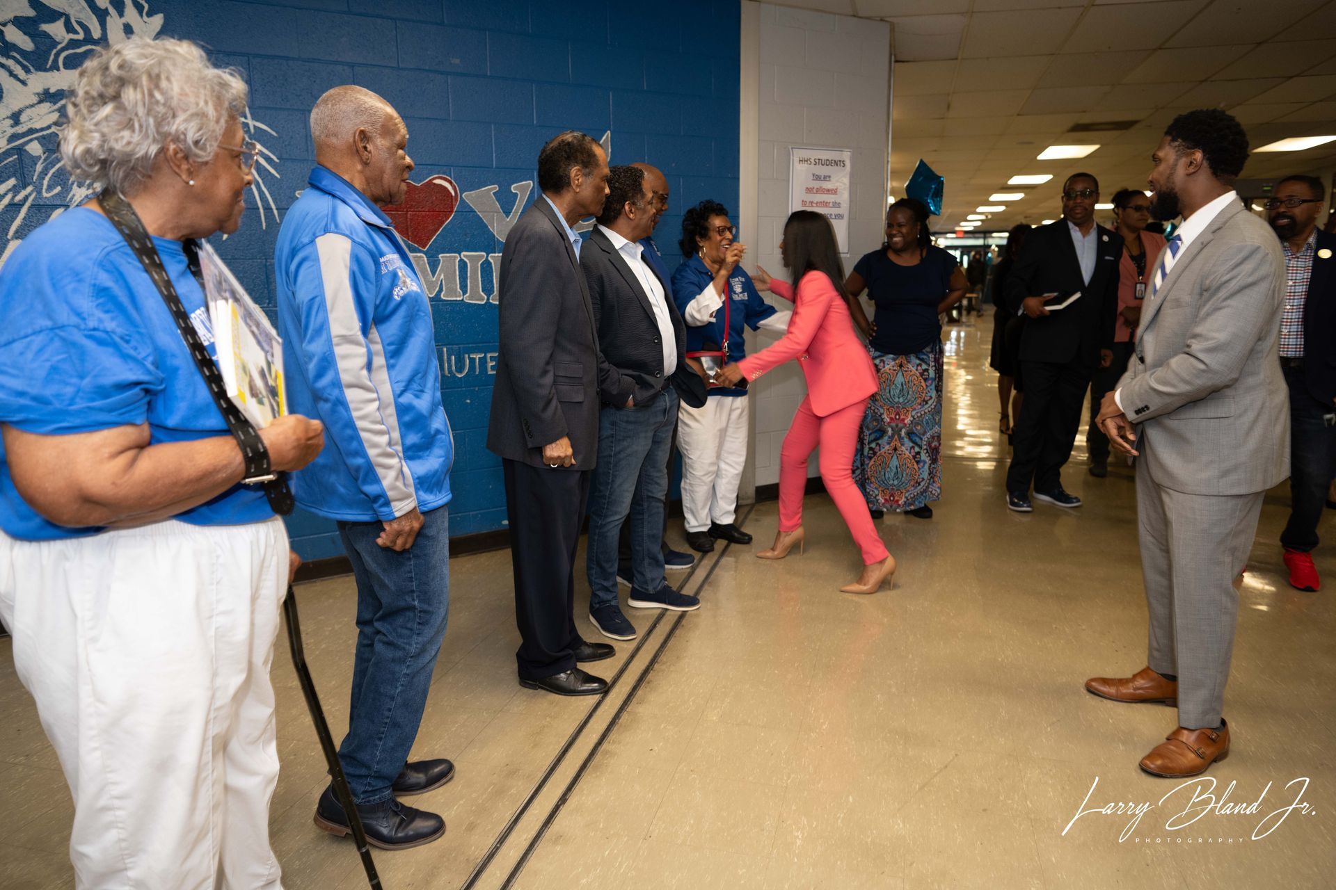 People in a hallway shaking hands. Woman in pink suit greets a group. Blue and gray tones.