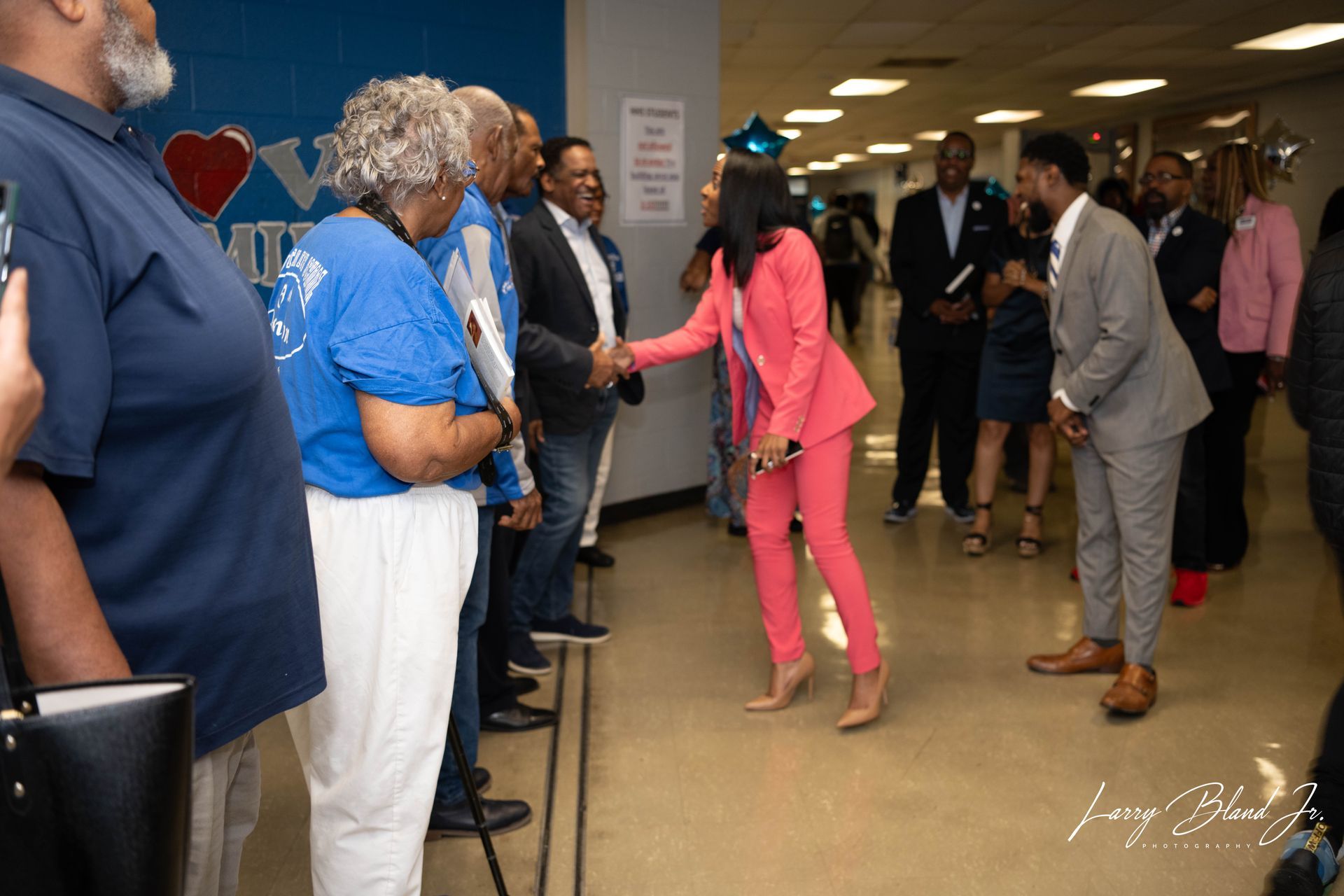 Woman in pink suit shakes hands with someone as others watch. Brightly lit hallway.