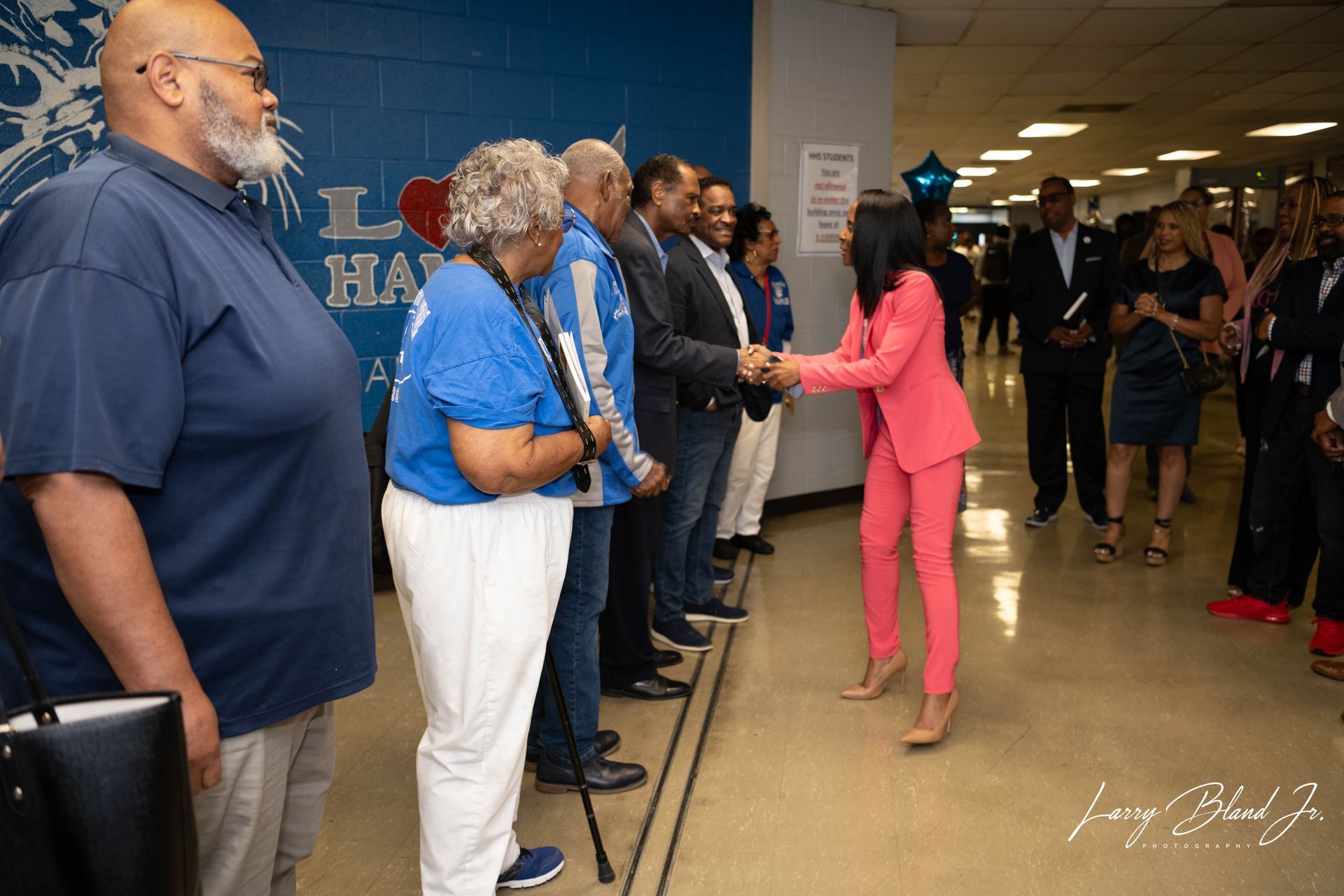 Woman in pink suit shakes hands with a line of people in a hallway; blue wall with a lion logo in the background.