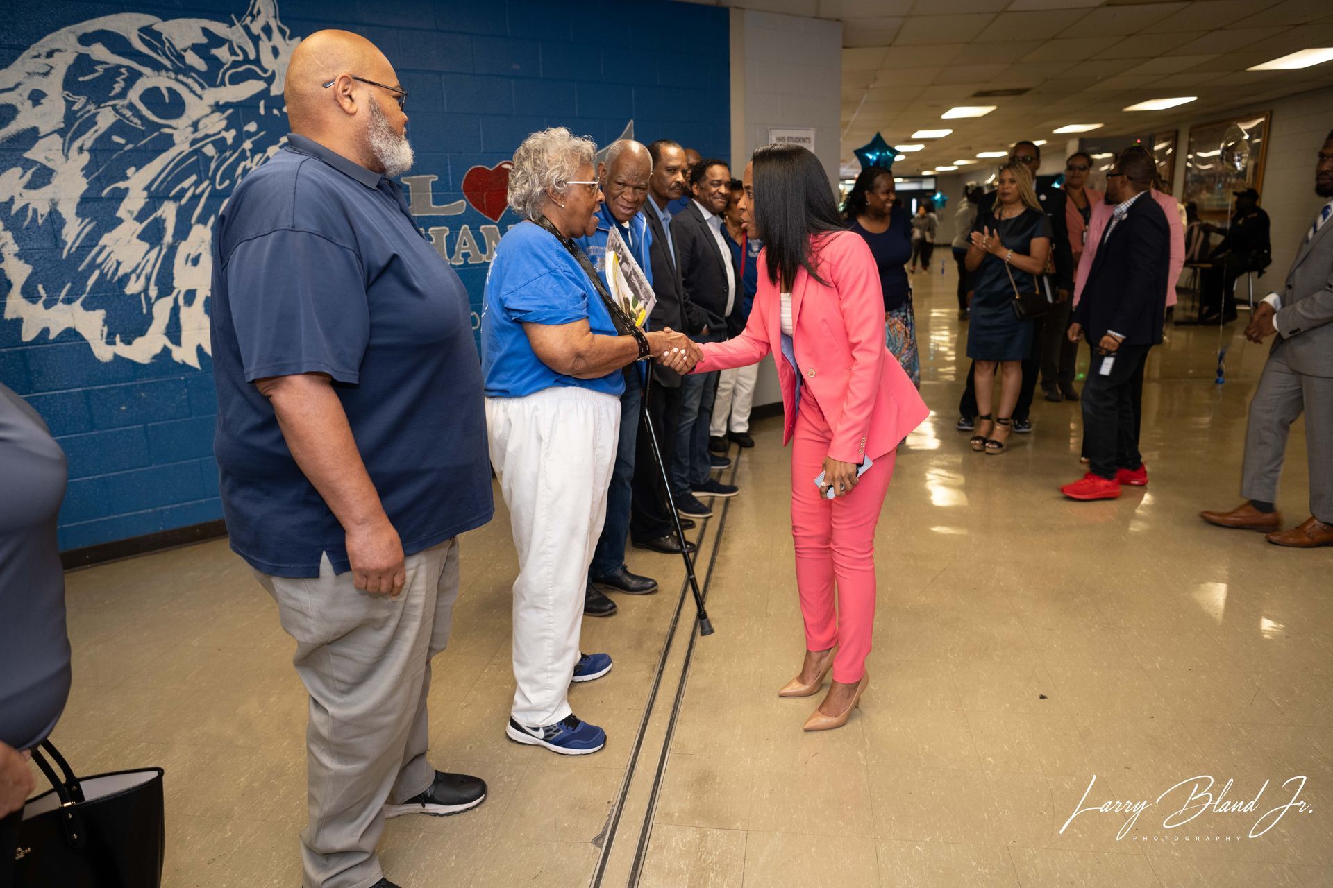 Woman in pink suit shakes hands with older person holding a plaque in school hallway.