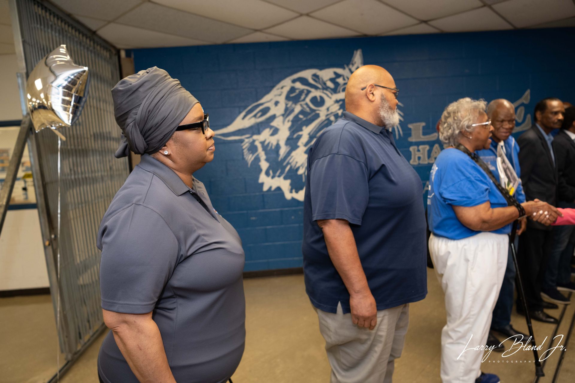 People standing in a room with a school logo on the wall. A woman wears a headwrap.