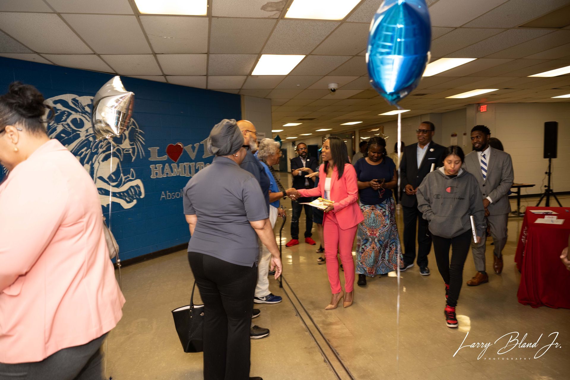 People gathered in a school hallway with balloons; woman in pink suit speaks to group.