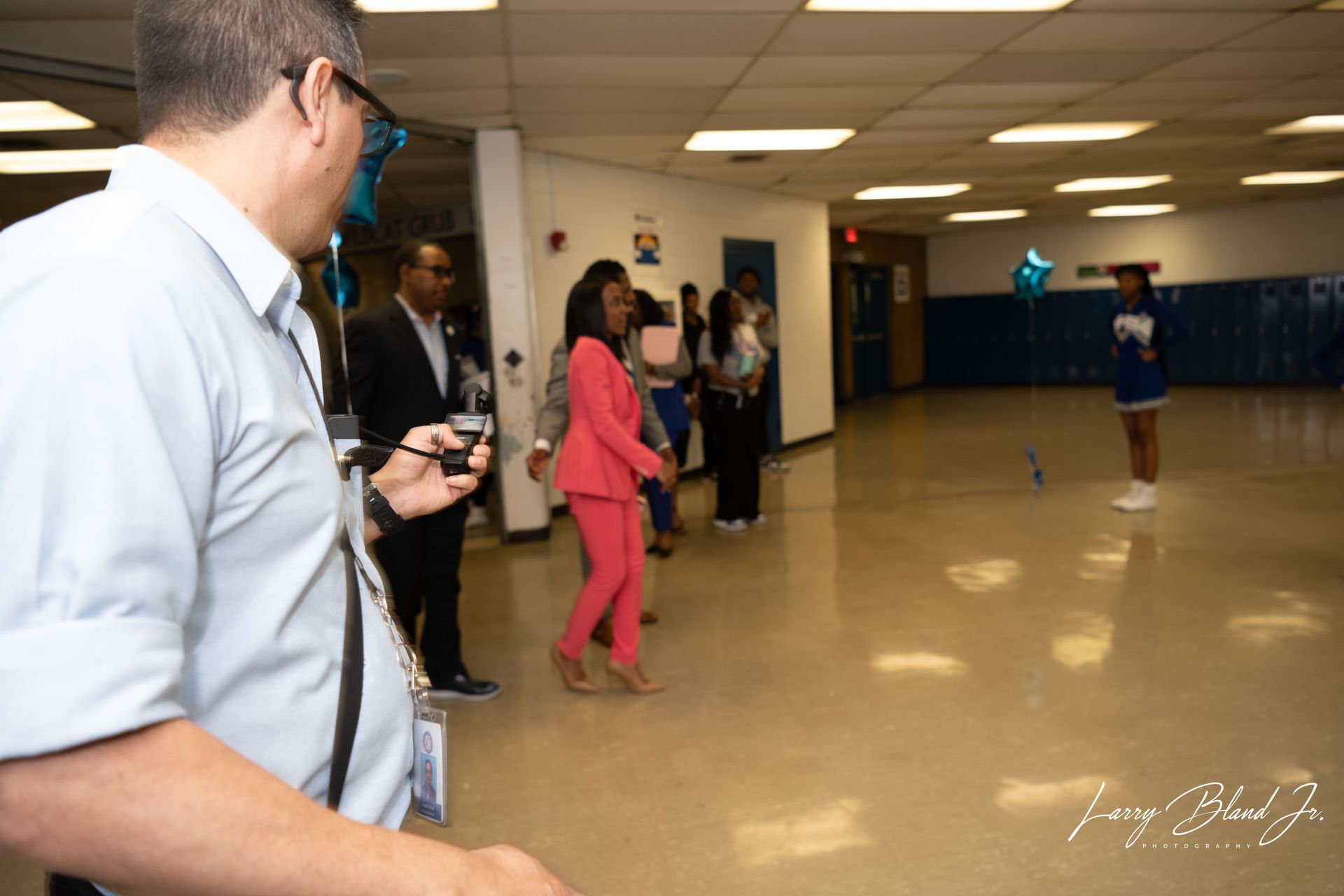 Person in pink suit walks in a hallway, people watching, a cheerleader with pom-poms.