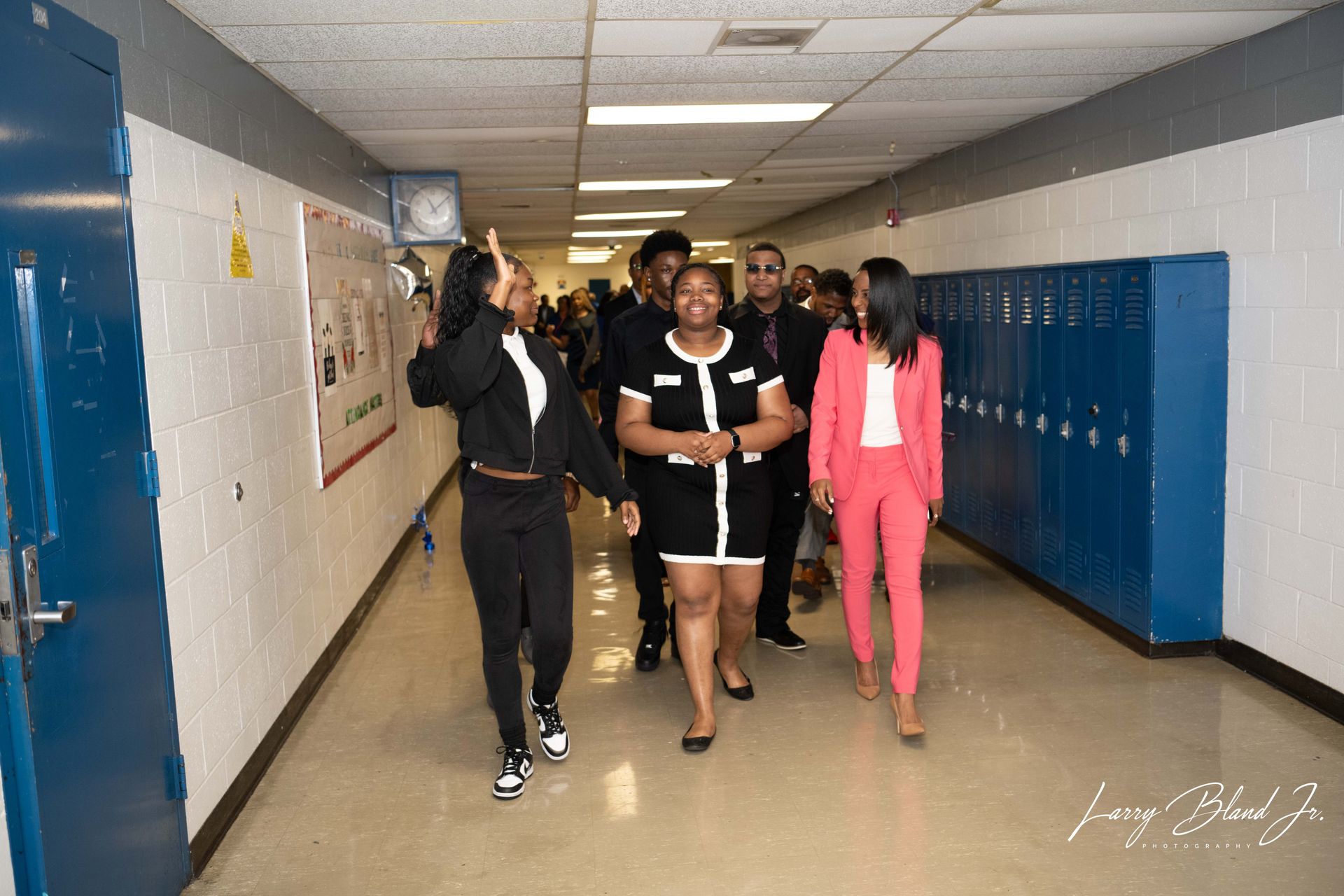Group of people walking down a school hallway. One in a pink suit, others in various outfits.