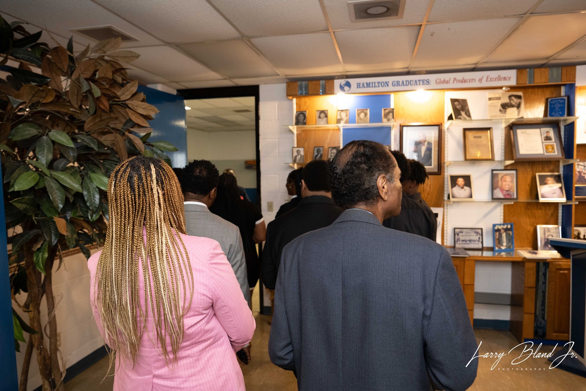 People entering a room, with a display of photos and awards on the wall.
