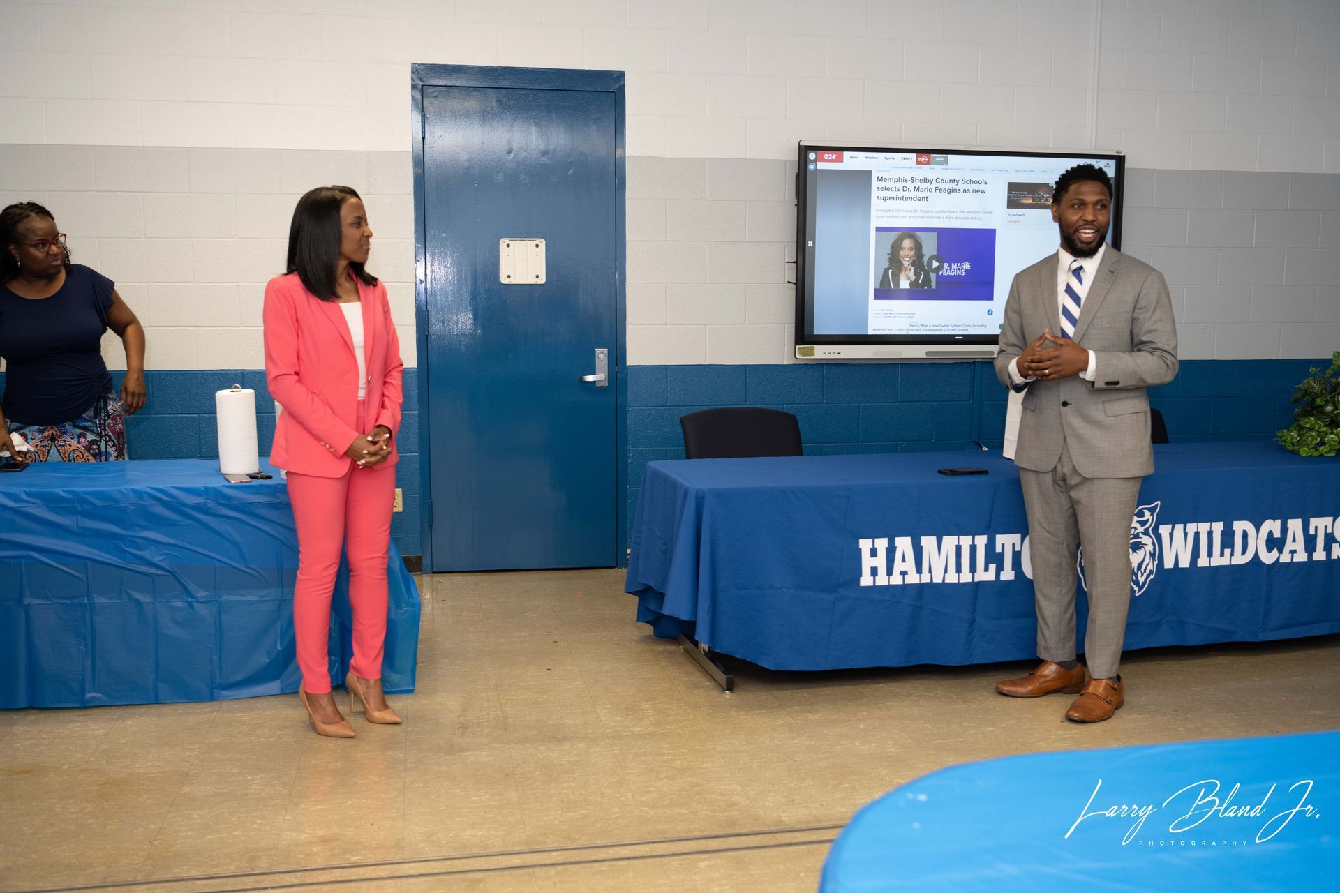People at a presentation: woman in pink suit, man in suit speaking, tables with 