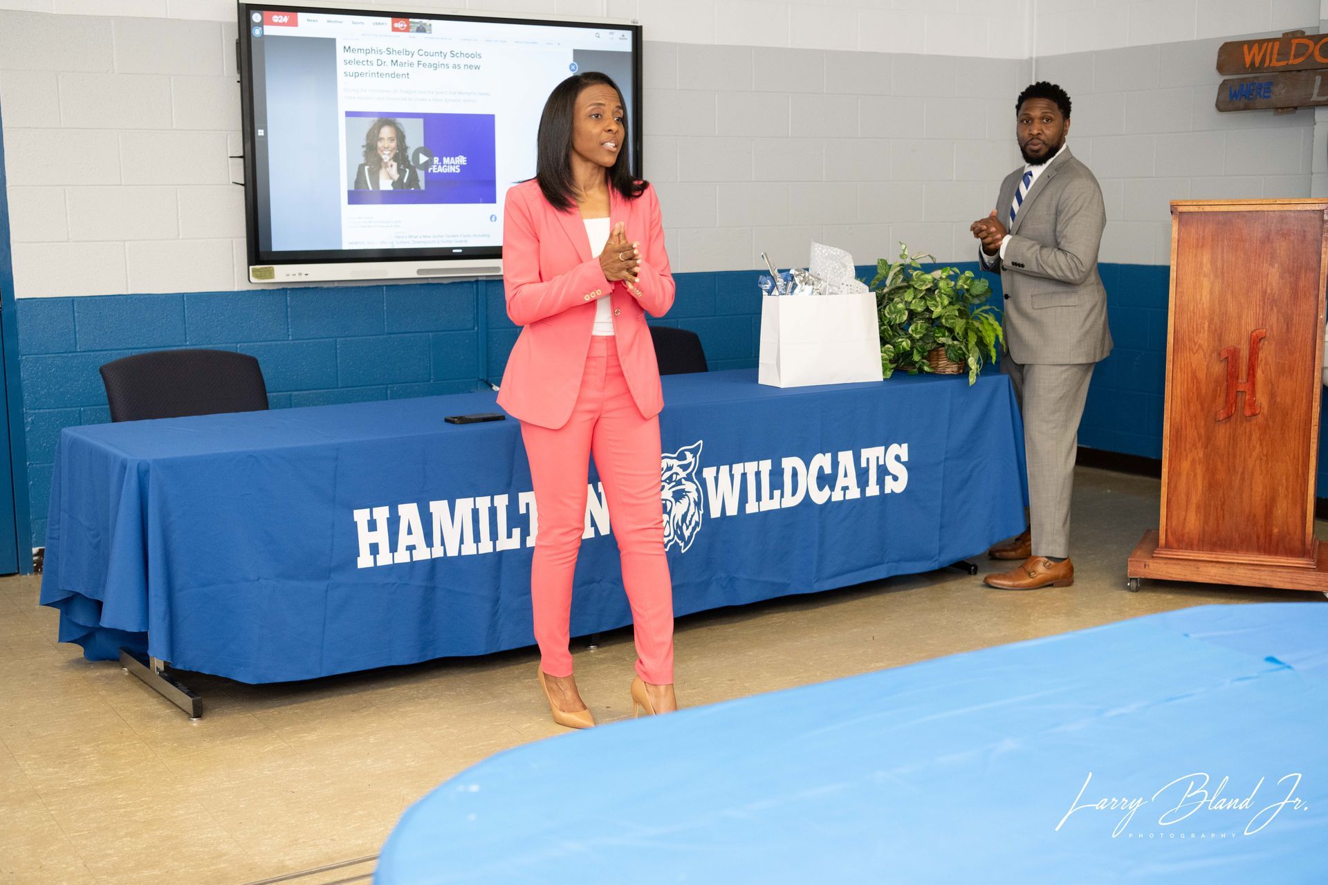 Woman in pink suit speaks at a podium. Man in suit stands beside table with 