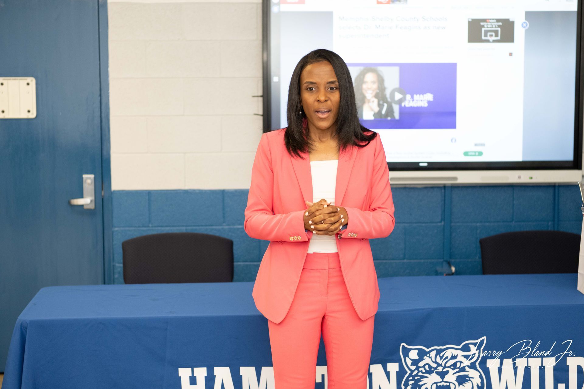 Woman in pink suit speaks at a table with a Hampton Wildcats sign, screen behind.
