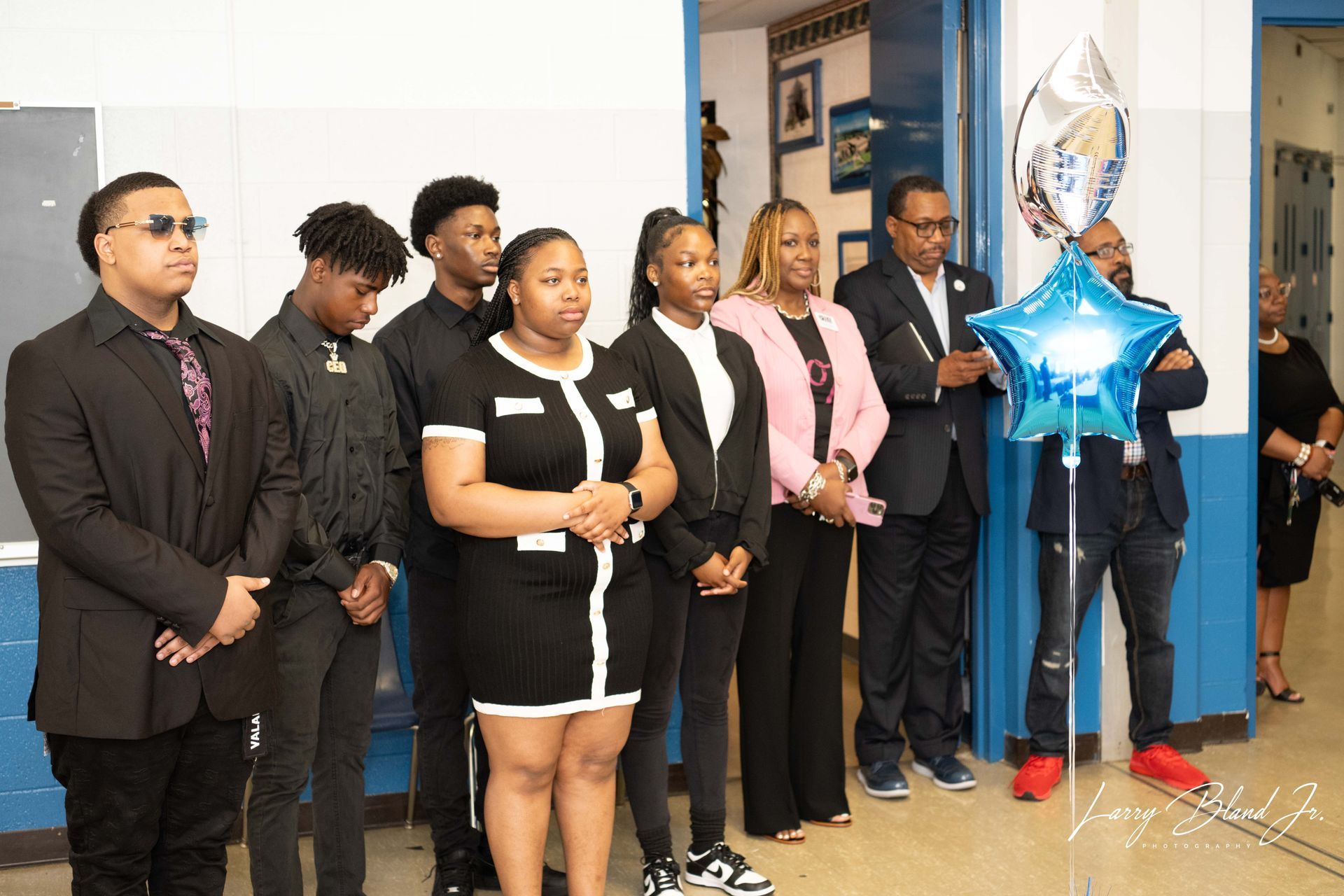 Group of people in a hallway, some wearing suits. One person holds a star-shaped balloon.