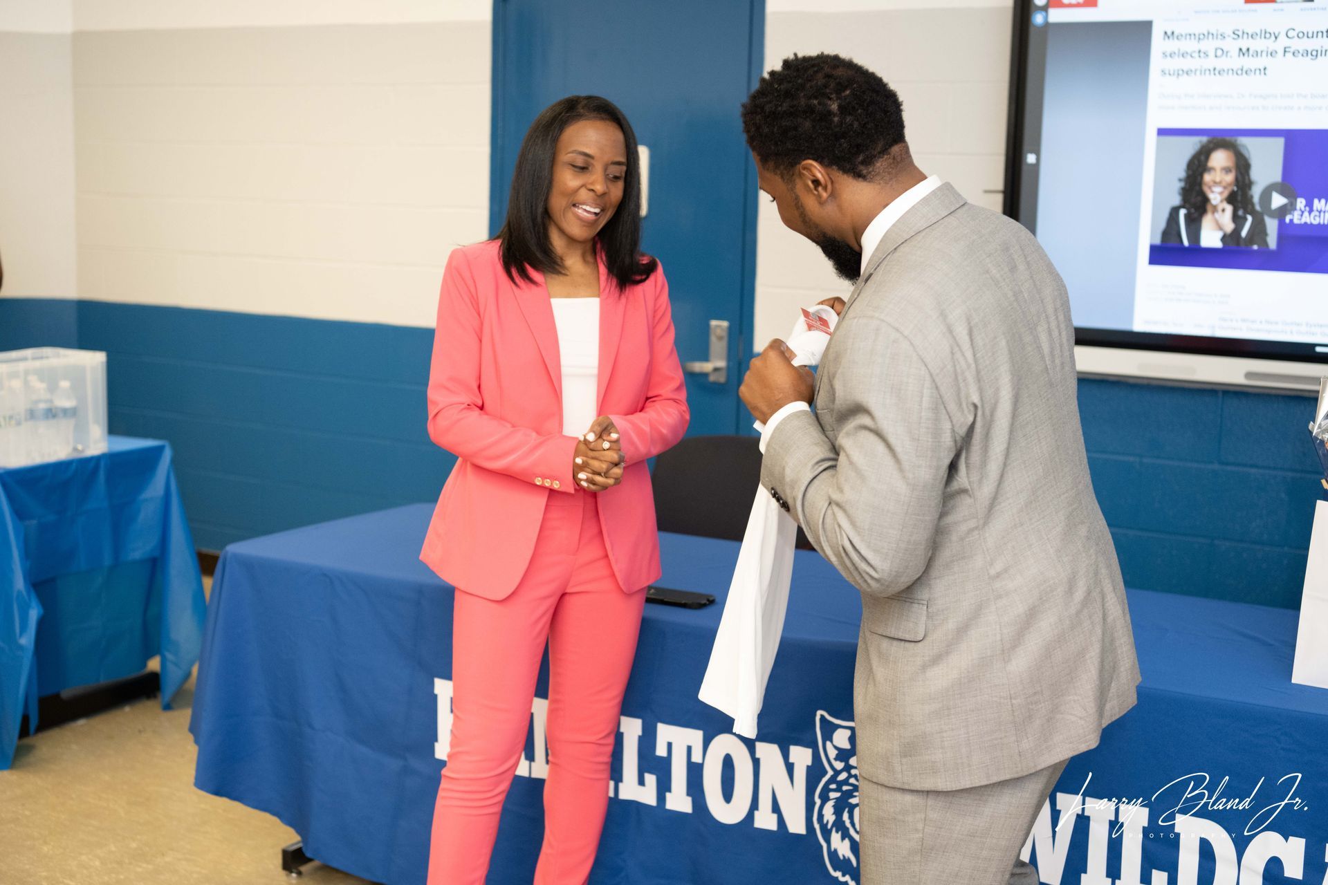 Woman in pink suit stands by table with man in gray suit. A presentation screen is behind them.