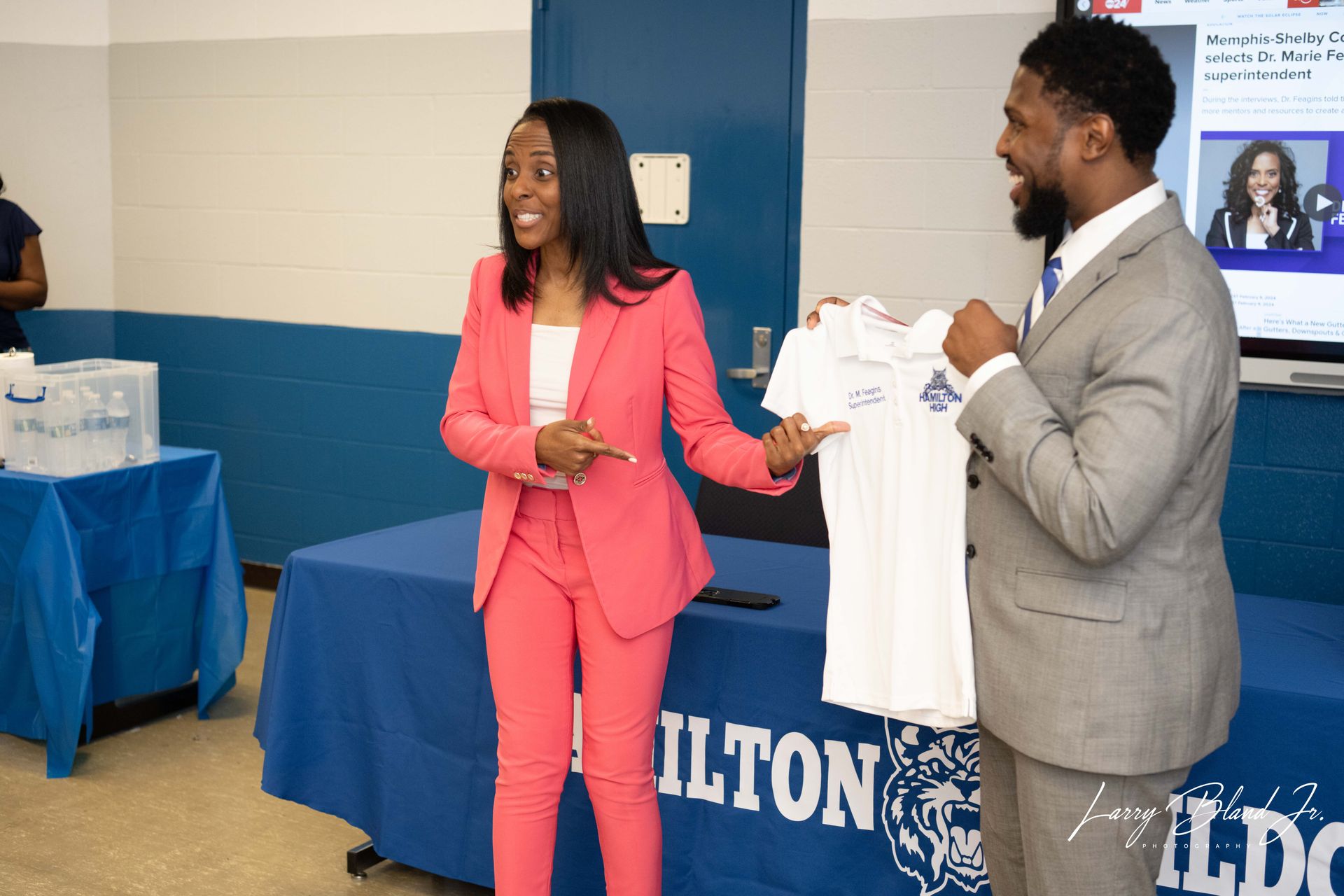 Woman in pink suit points, man in suit holds a shirt, both at an event with a blue table.