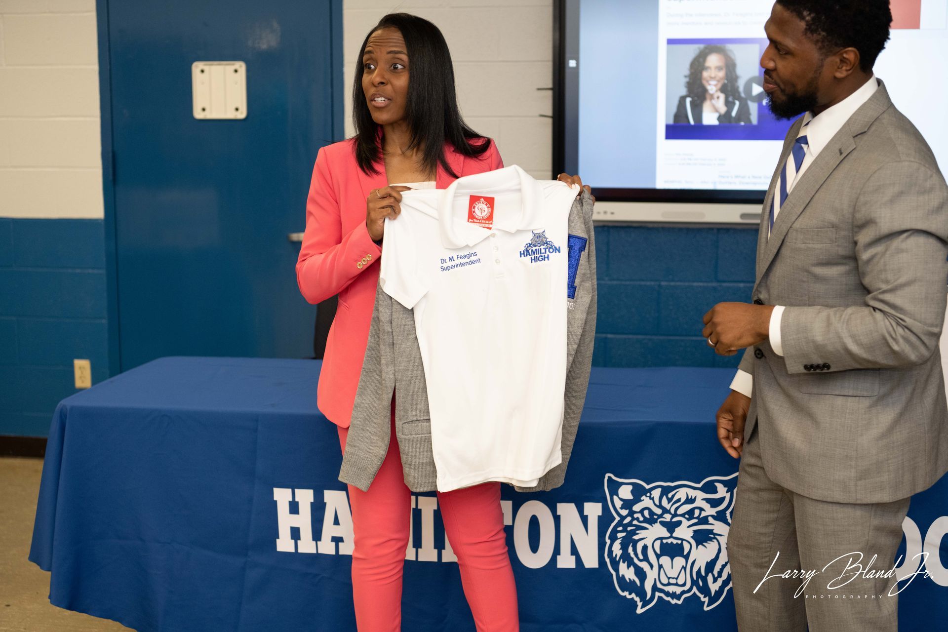 Woman holding up a white shirt with logo next to a man in a suit at a table with 