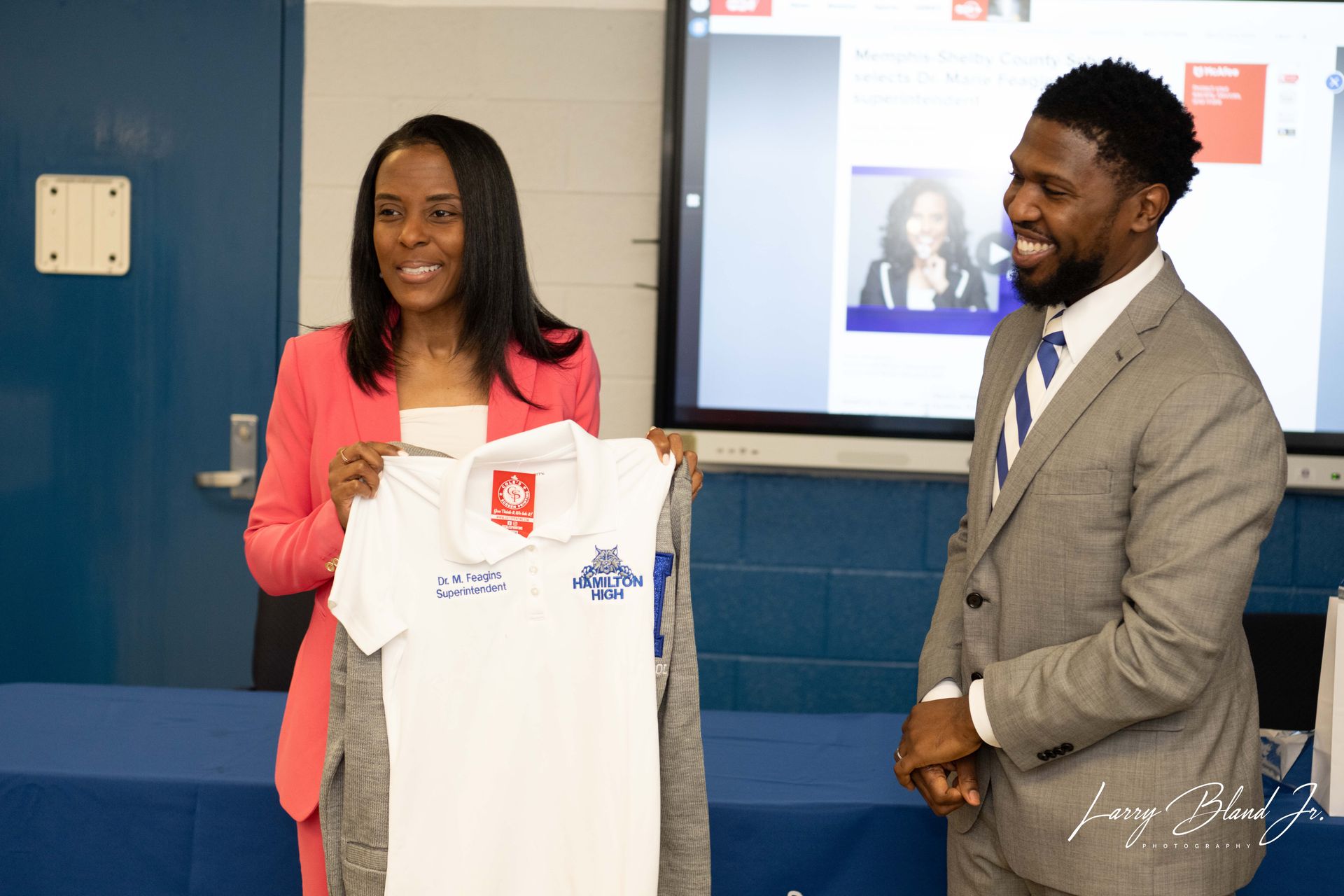 Woman in pink blazer holding white shirt, smiling man in suit looks on. In a room, presentation screen in background.