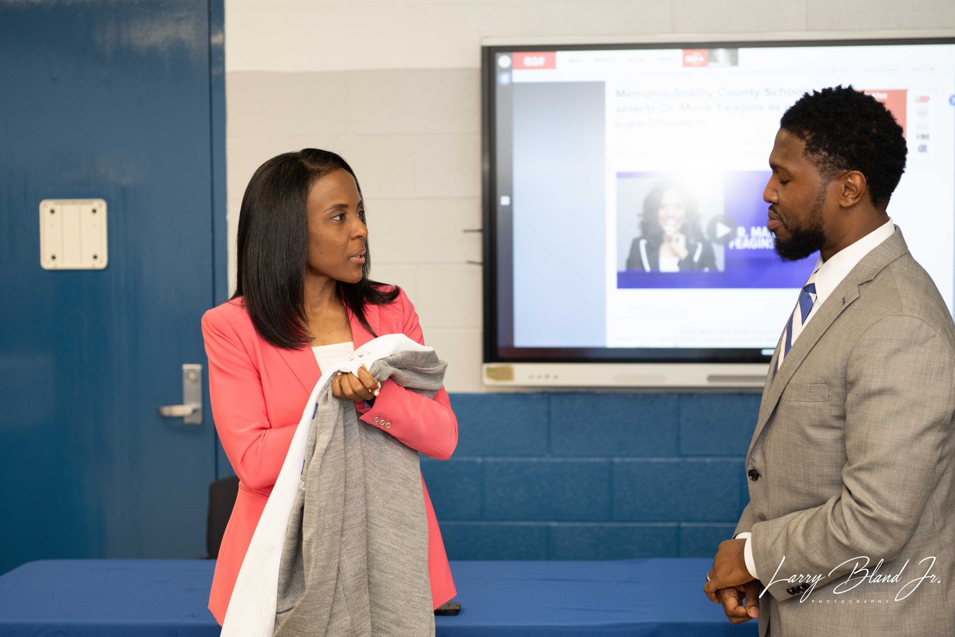 Woman in pink jacket holds a coat, speaking to a man in a suit, in front of a screen and blue wall.