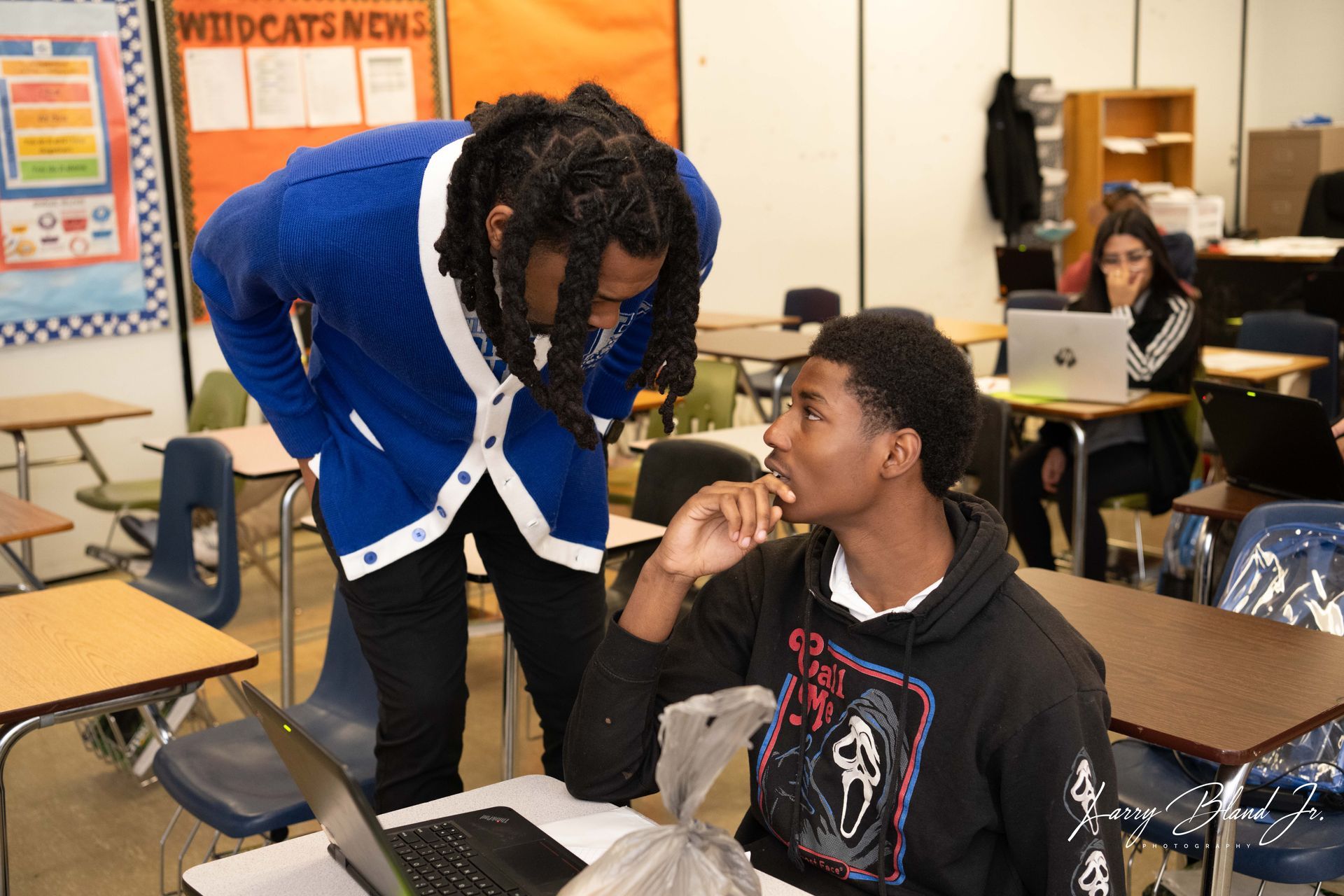 Two students in classroom, one leaning over another working on a laptop. Blue cardigan, black hoodie.