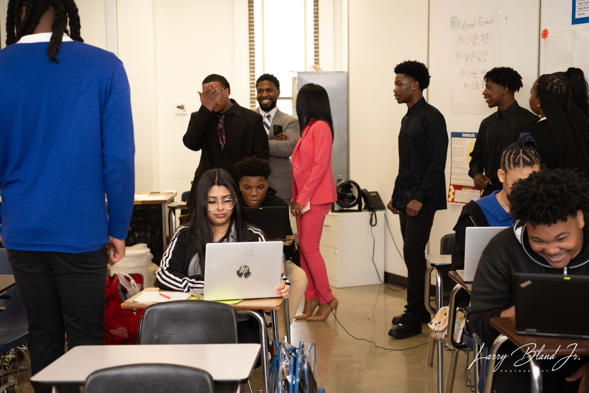 Classroom with students on laptops, presenters, and spectators; woman in pink suit speaks.
