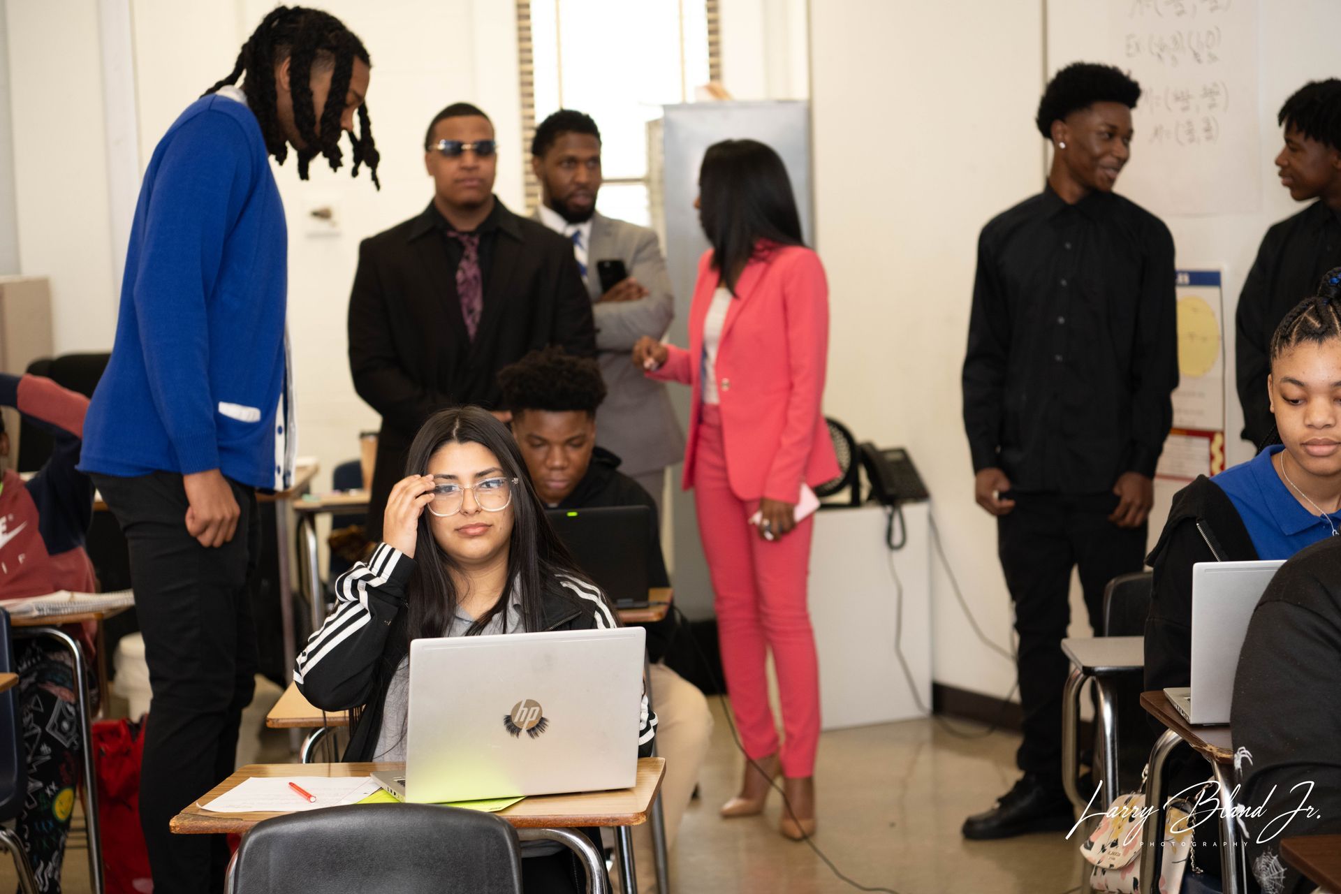 Woman in red suit speaking to students in a classroom. Students are at desks with laptops, others standing.