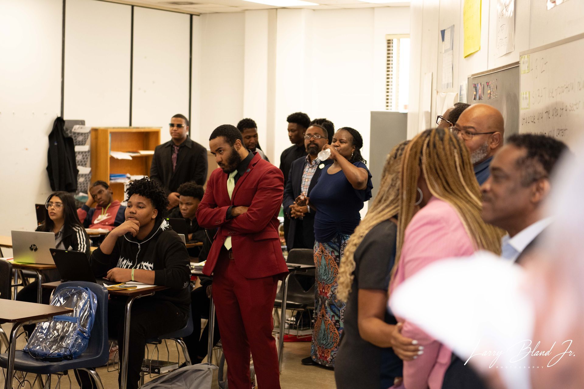 People in a classroom, some standing and some seated. A man in a red suit stands out.