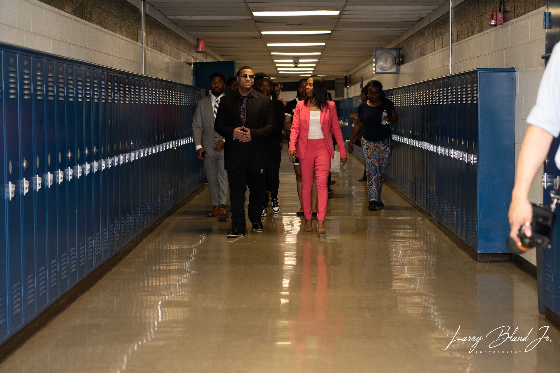 People walking down a school hallway lined with blue lockers. One person wears a pink suit.