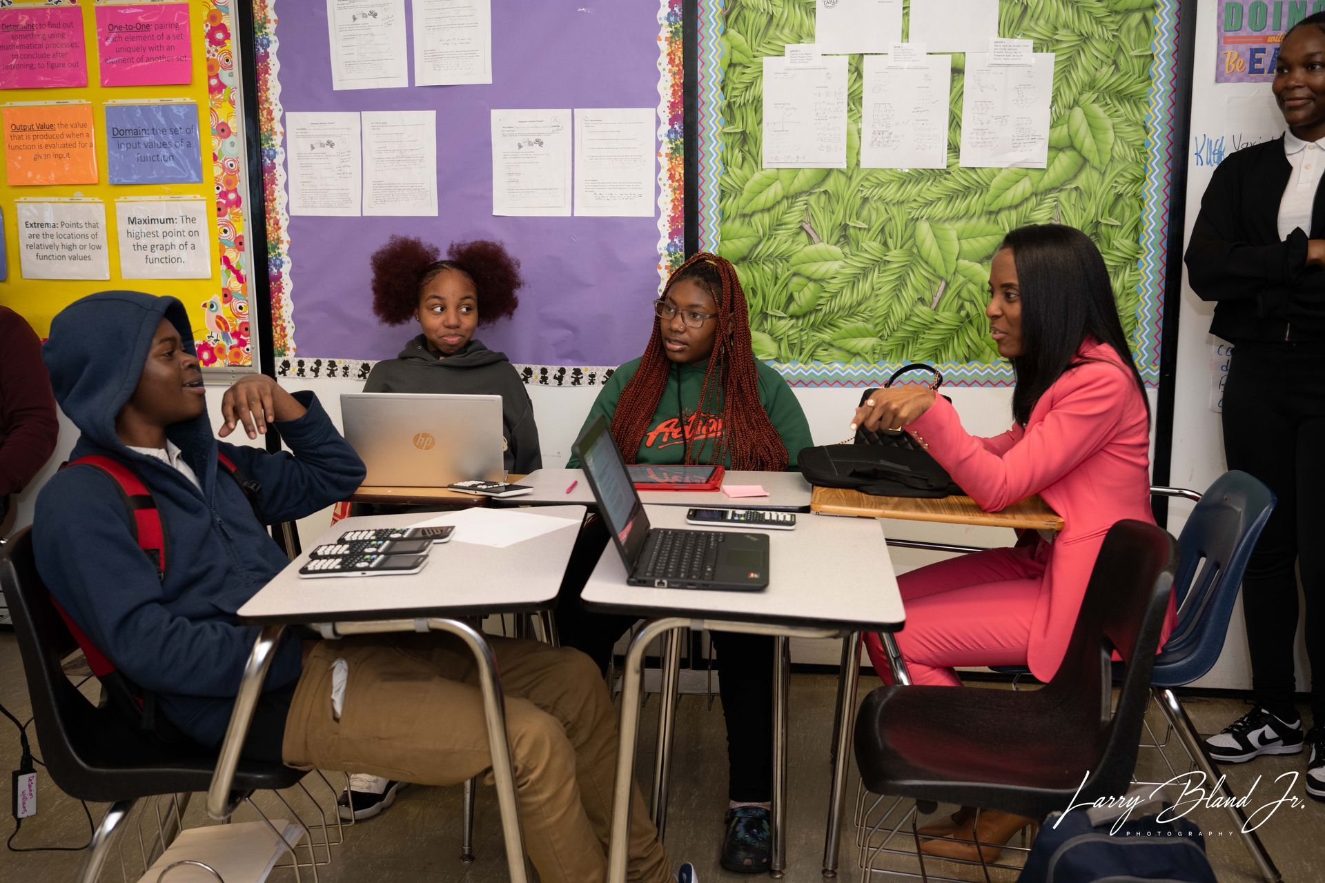 Students in classroom at desks with laptops, listening to a woman in a pink suit; another person looks on.