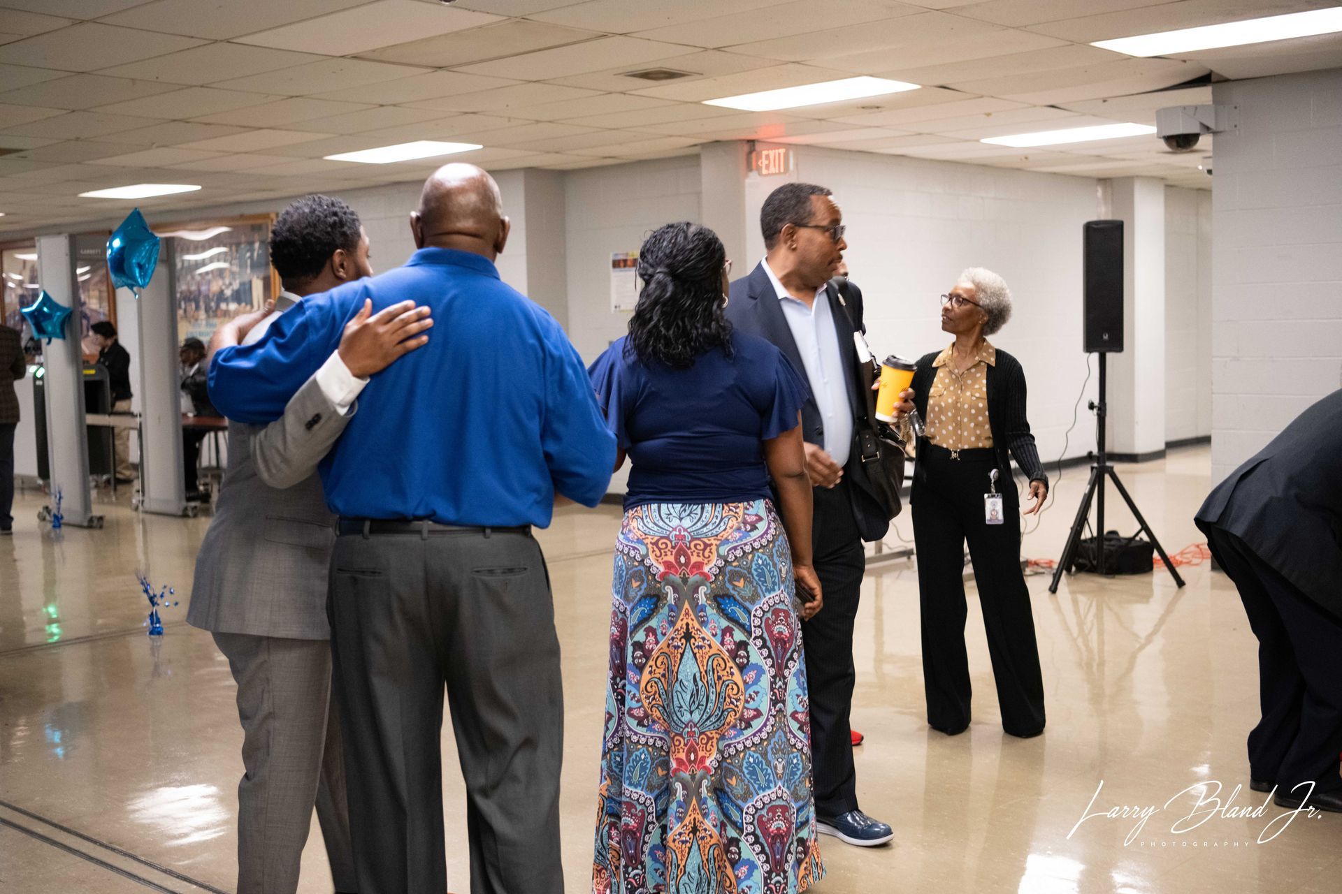 People hugging and talking in a hallway, possibly at an event. Some are wearing suits and dresses.